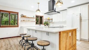 A modern kitchen with a large island, wooden stools, pendant lighting, and white cabinetry.