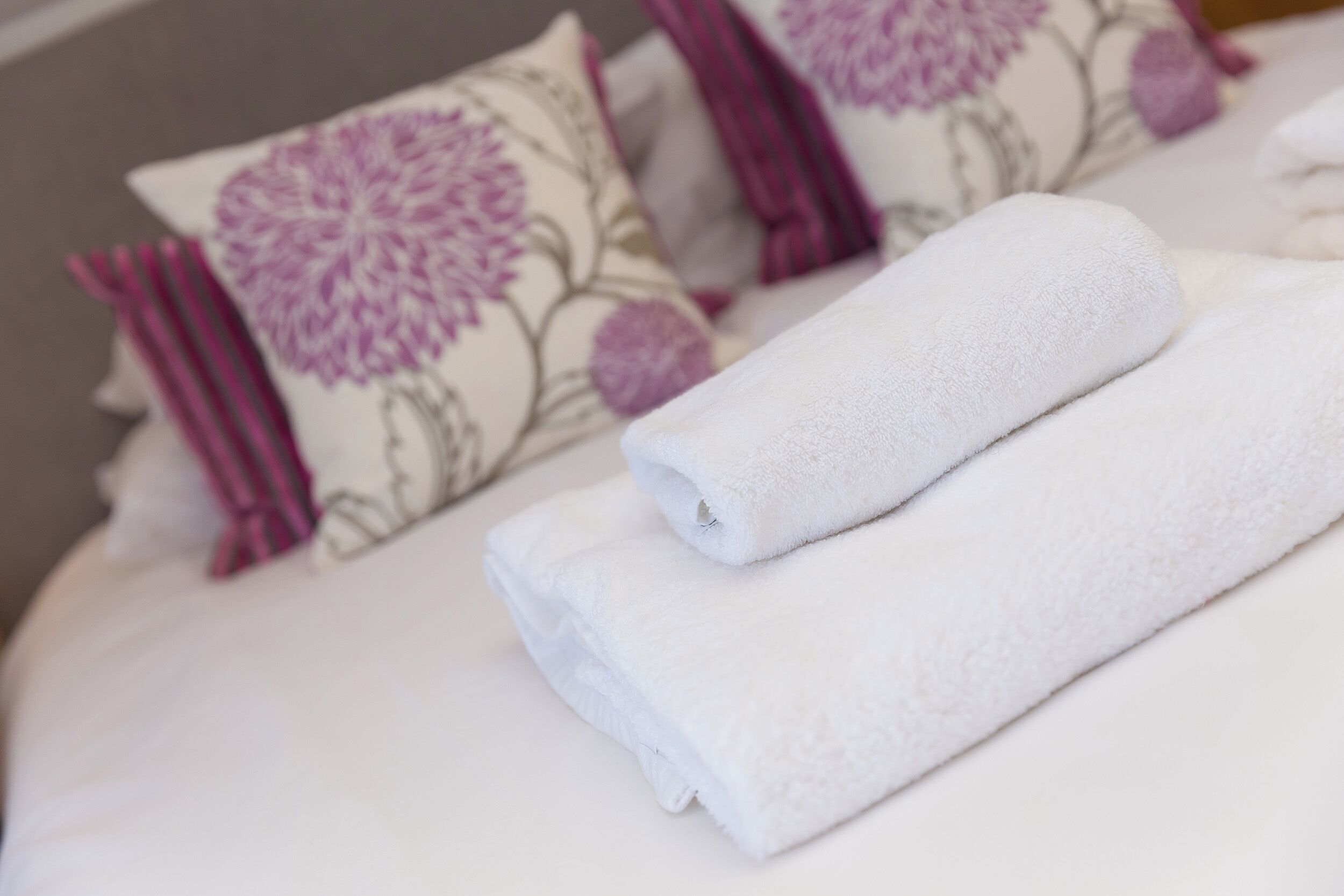 Una cama bien hecha con sábanas blancas y almohadones con motivos florales púrpuras.