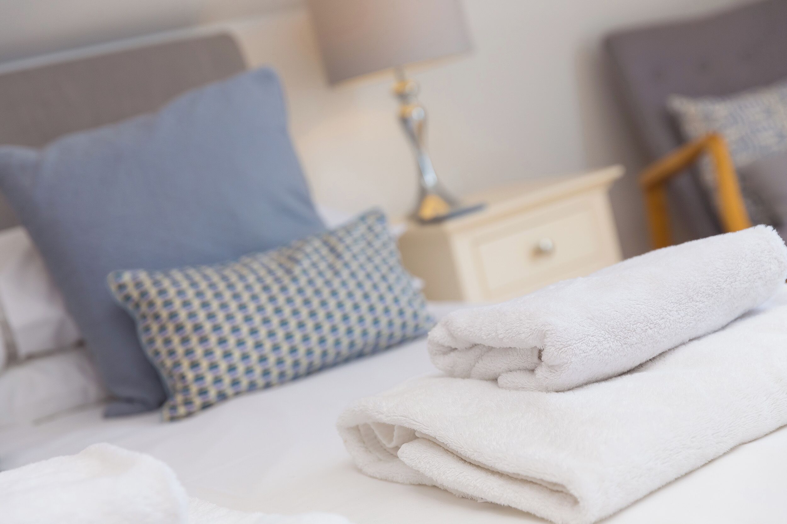 A neatly made bed with white and patterned pillows, a folded white towel, and a bedside table with a lamp.