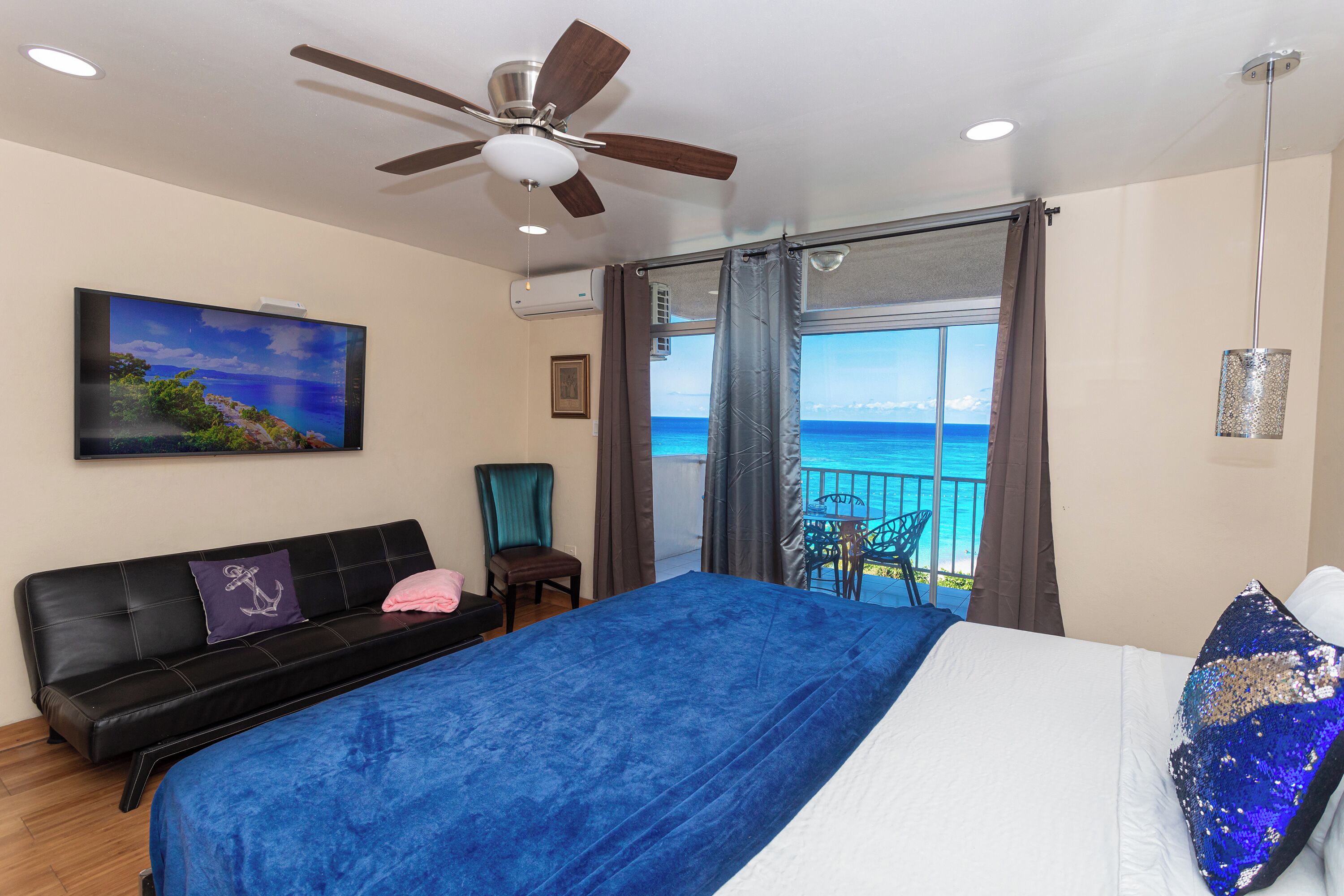 A bedroom with a bed, a ceiling fan, a TV, a balcony with a view of the sea, and a chair.