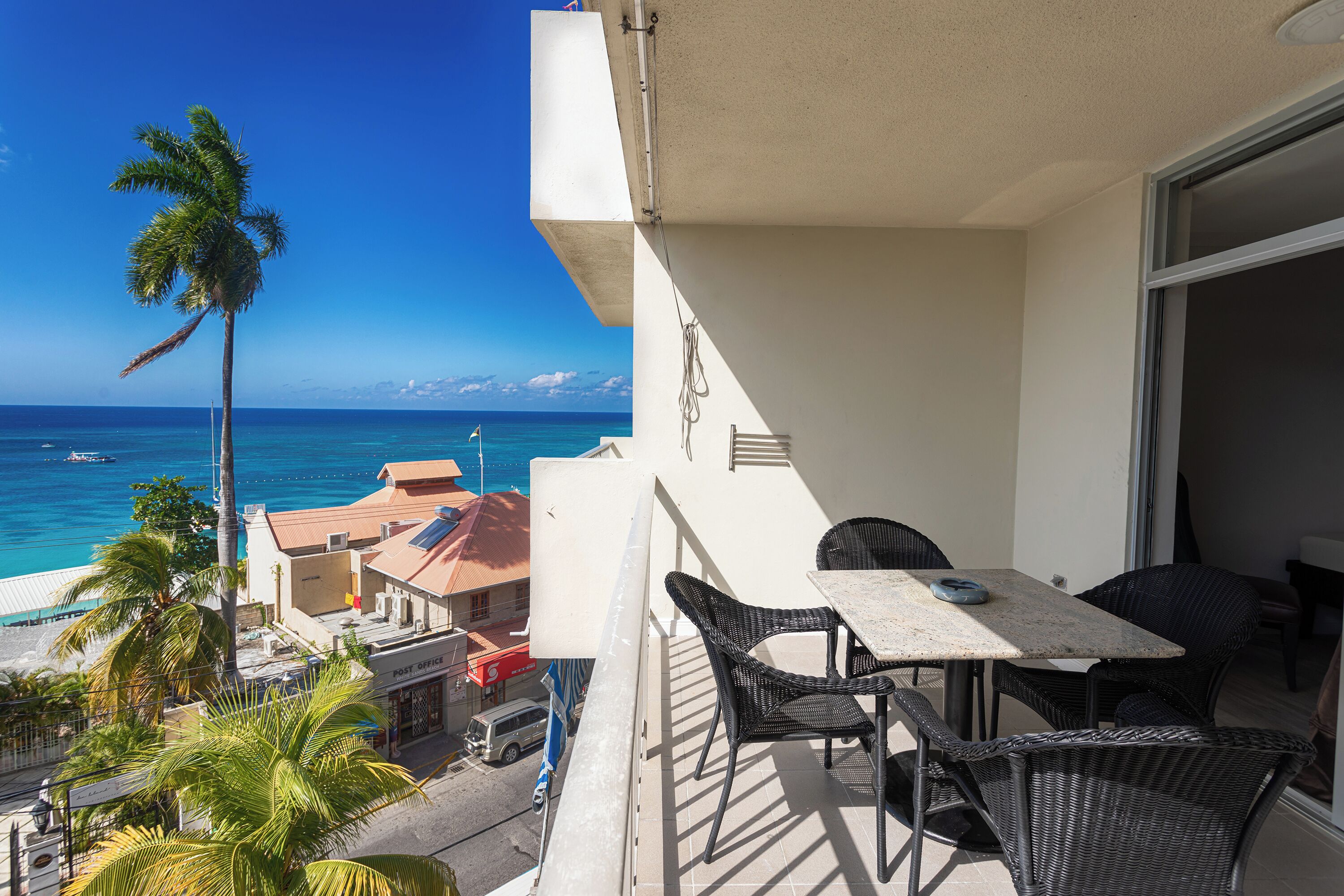 A balcony with a table and chairs, overlooking a beach with palm trees and a clear blue sky.