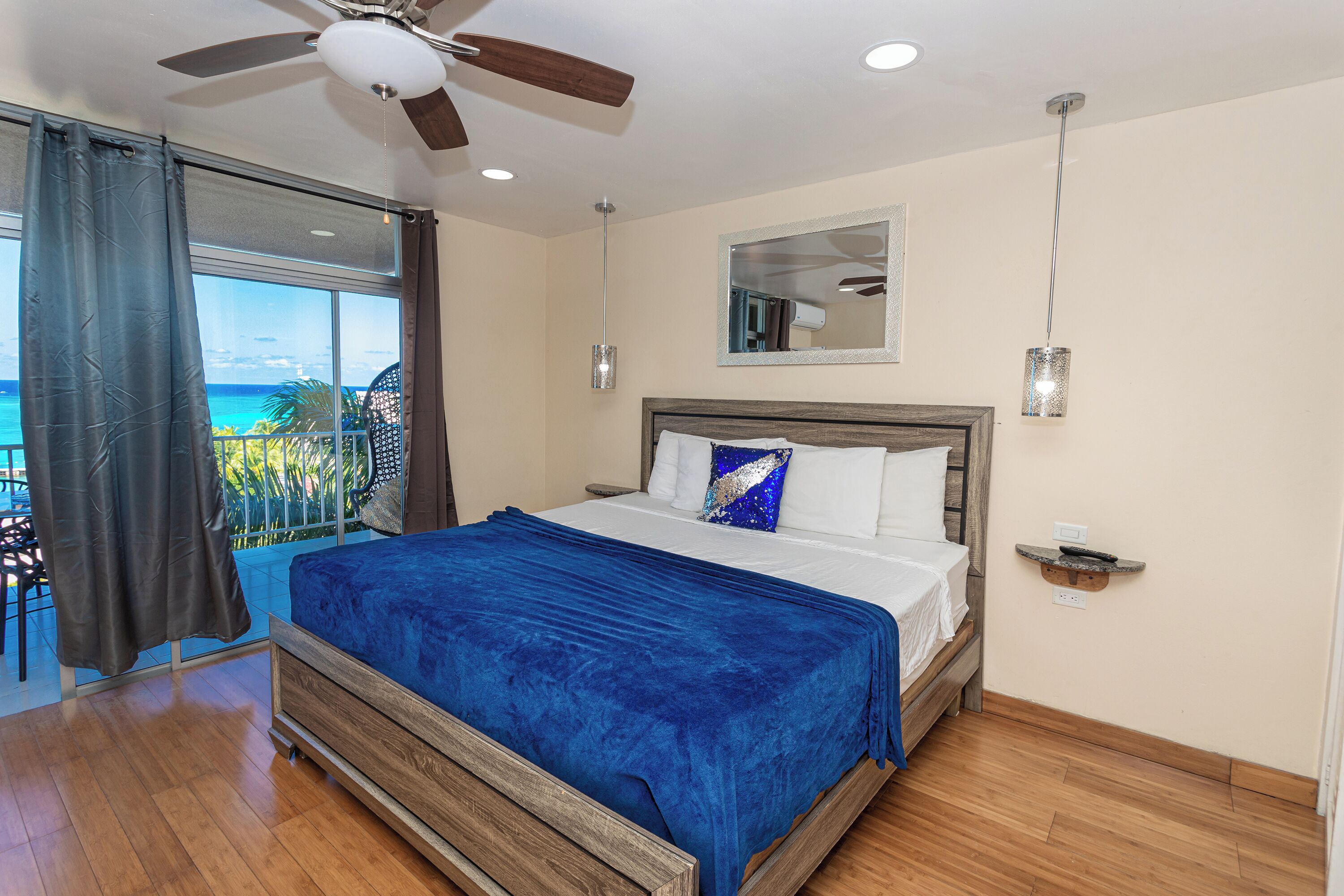 A bedroom with a bed, a ceiling fan, a view of the ocean, and a balcony with a chair.
