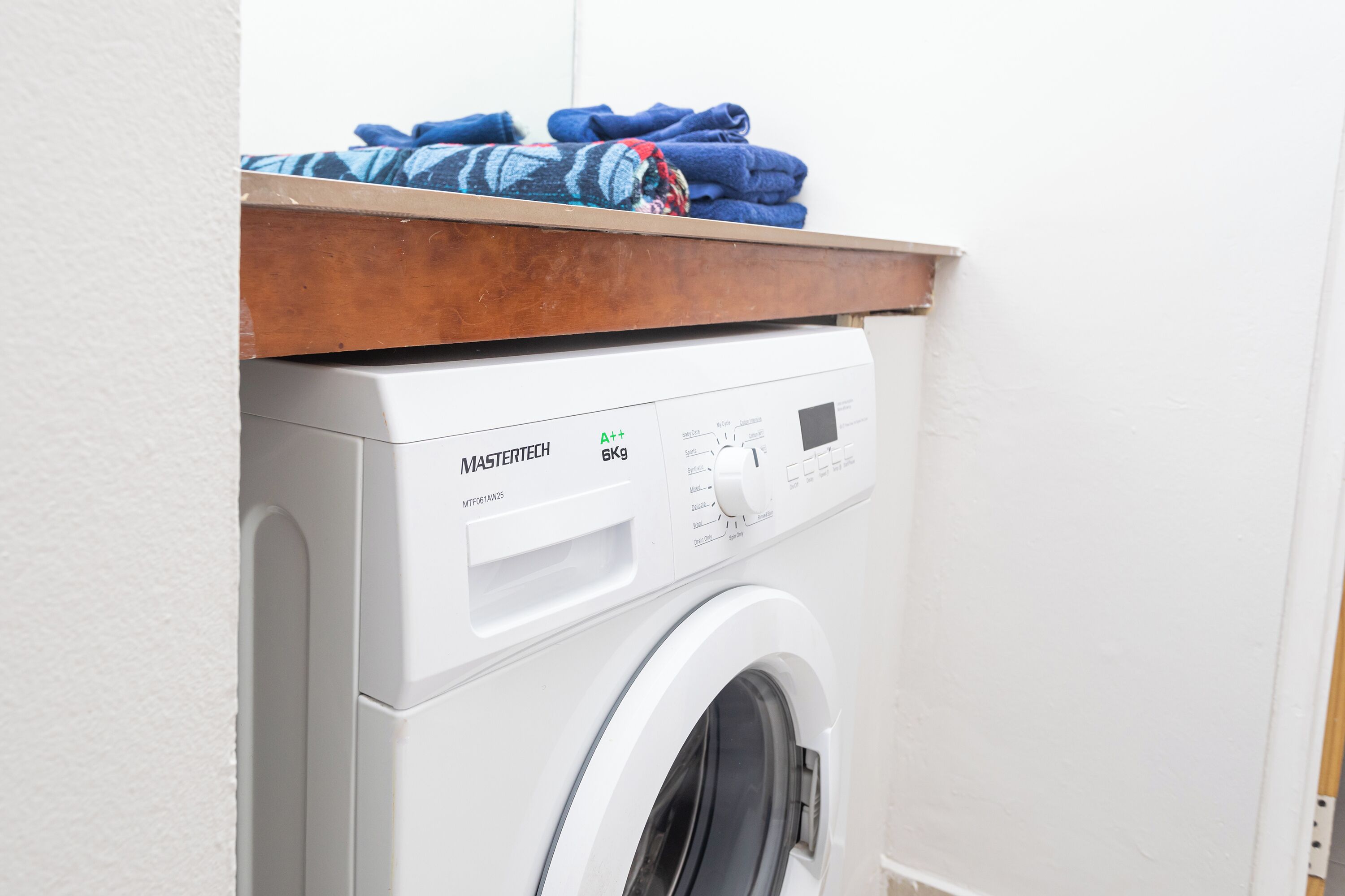 A front-loading washing machine with a control panel, a wooden shelf above it, and a white wall in the background.