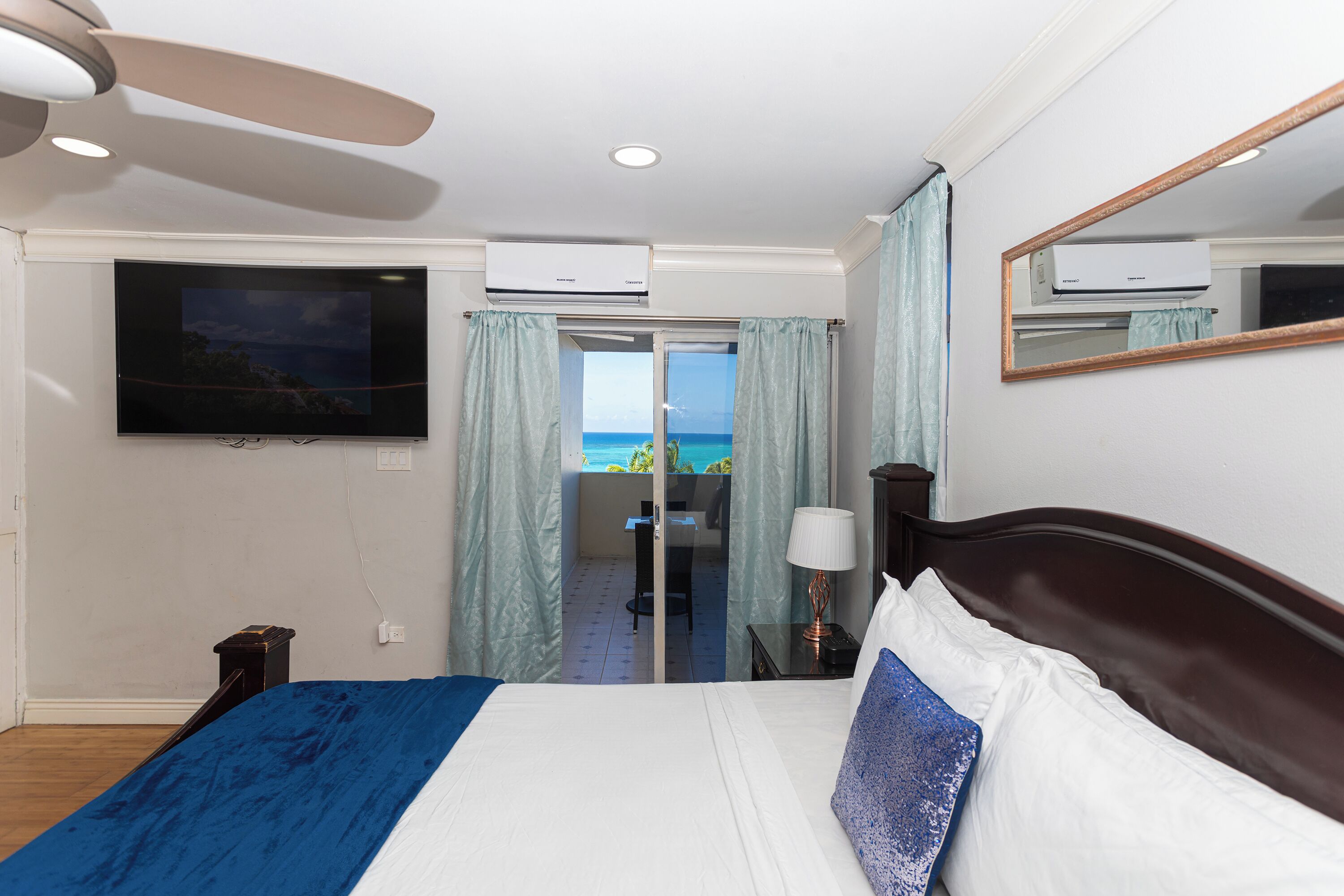 A hotel room with a bed, a TV, an air conditioner, a ceiling fan, and a view of the ocean through a sliding door.