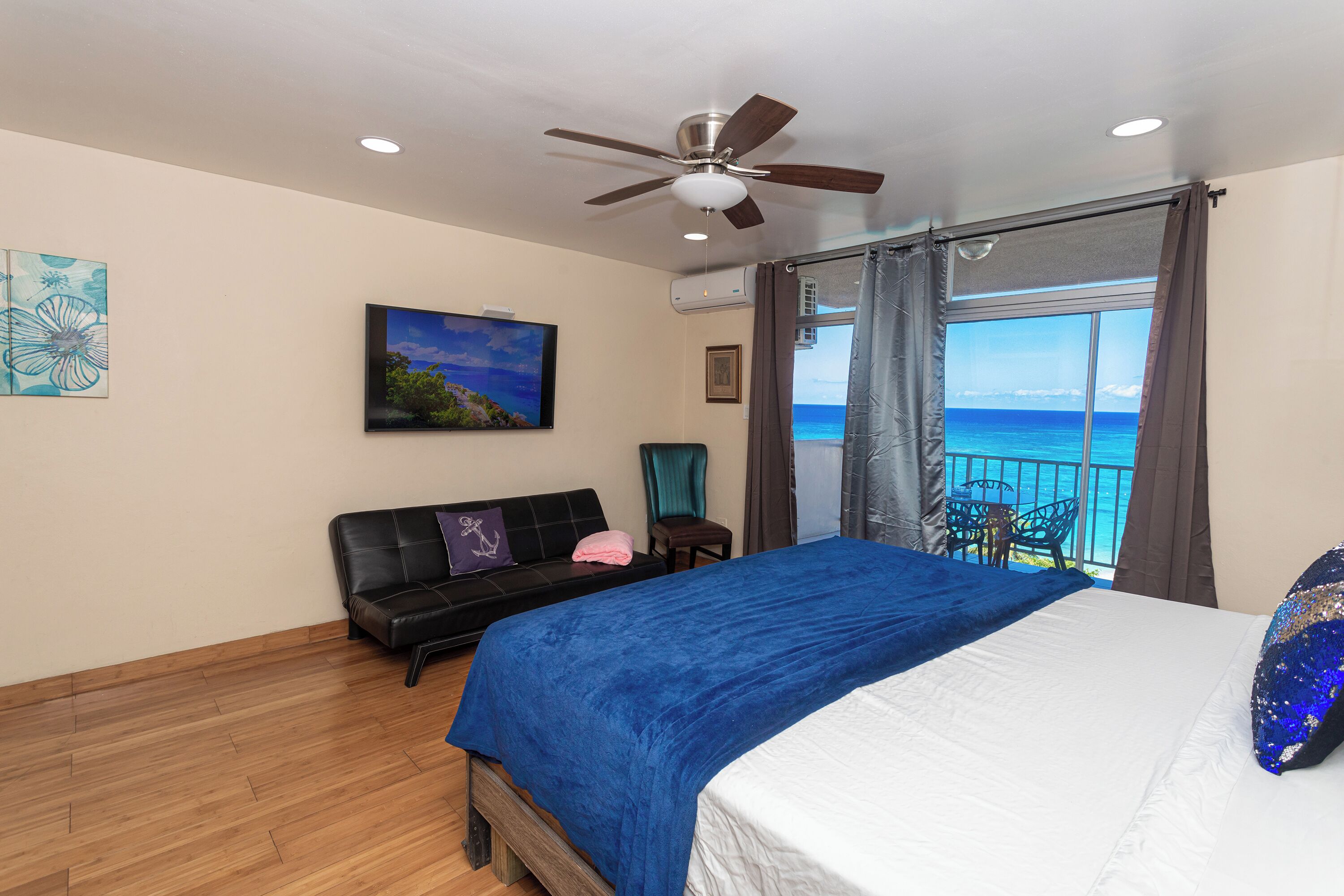 A bedroom with a bed, a ceiling fan, a view of the ocean, a balcony with a table and chairs, and a painting on the wall.