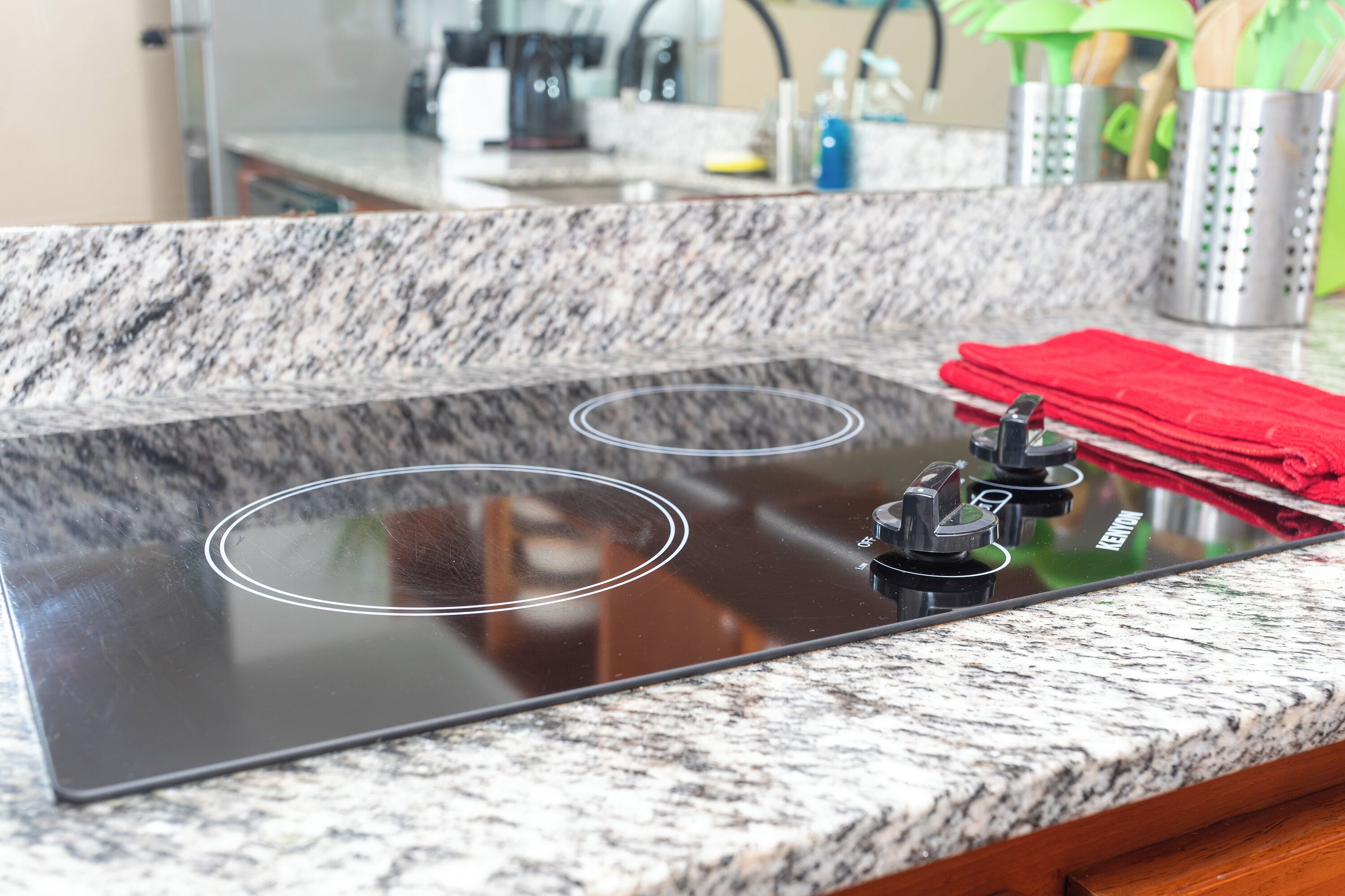 Une cuisine dotée d’une plaque de cuisson à induction noire, de plans de travail en granit et d’un torchon rouge posé sur le plan de travail.