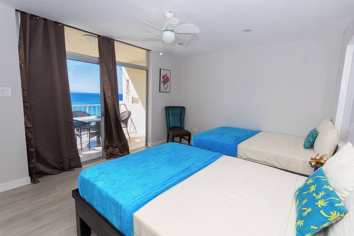 A hotel room with two beds, a ceiling fan, a chair, and a view of the sea.