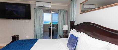 Quarto de hotel com cama, televisão na parede, aparelho de ar condicionado, janela com cortinas e vista para o mar.