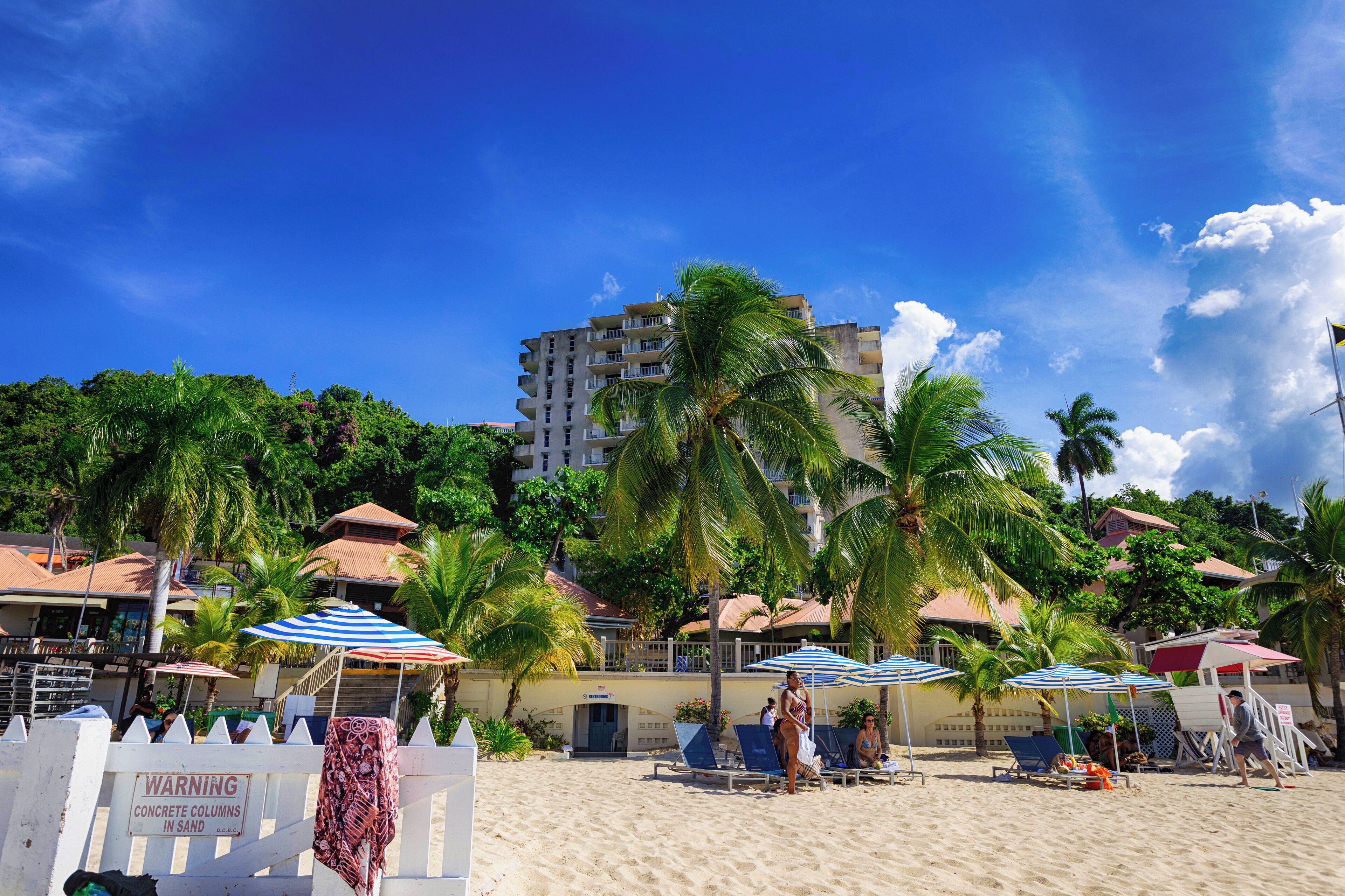 A beach with palm trees, beach chairs, and umbrellas. There is a building with a sign that reads 'WARNING CONCRETE COLUMBS IN SAND.'
