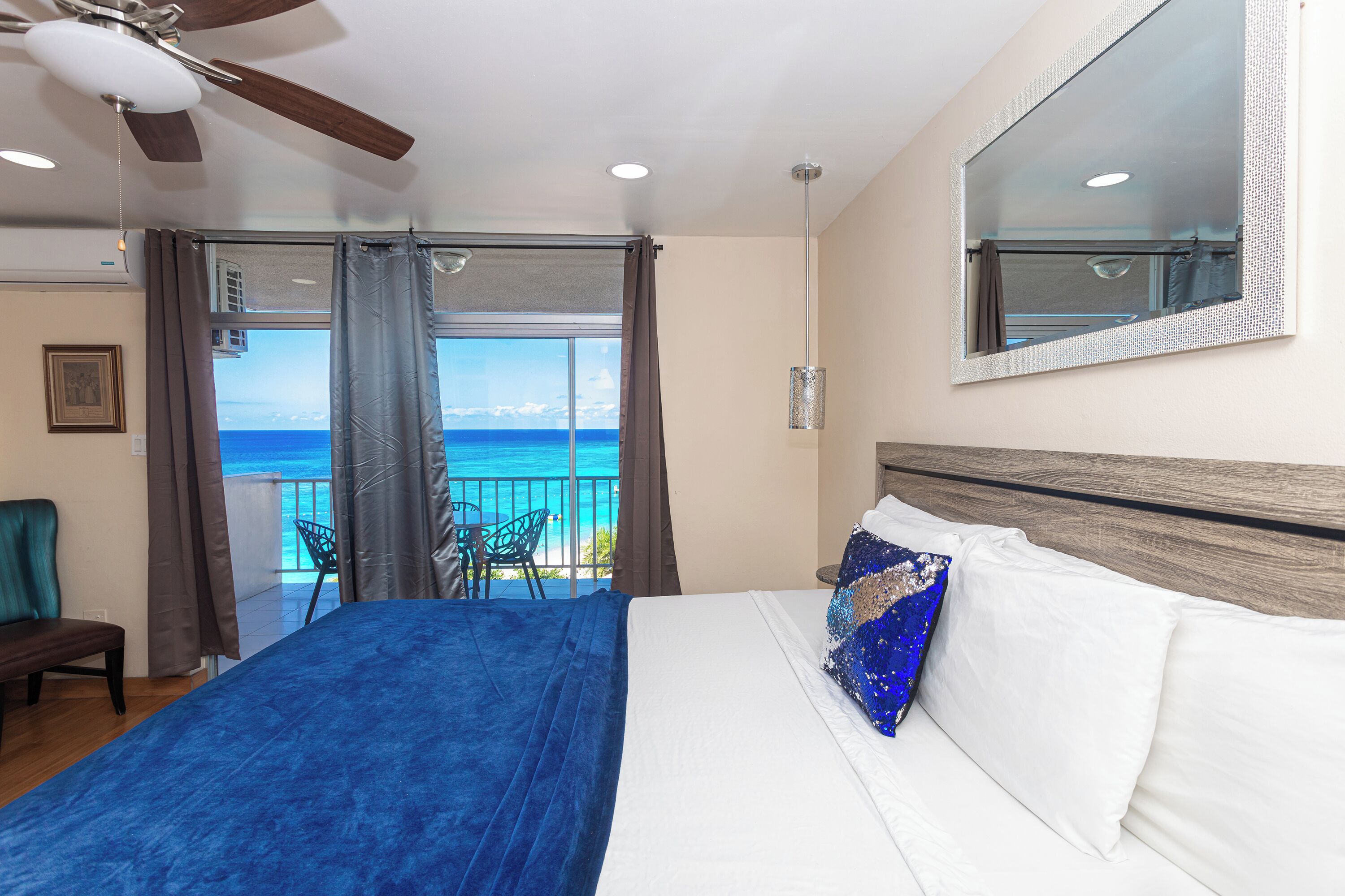 Quarto com cama, ventoinha no teto, janela com cortinas, varanda com cadeira e vista para o mar.