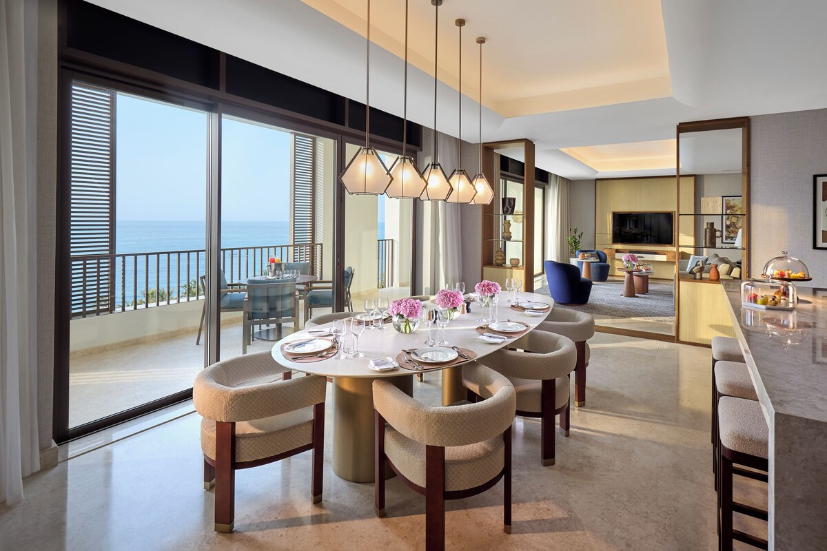 A dining area with a large round table, multiple chairs, and a view of the sea through large windows.