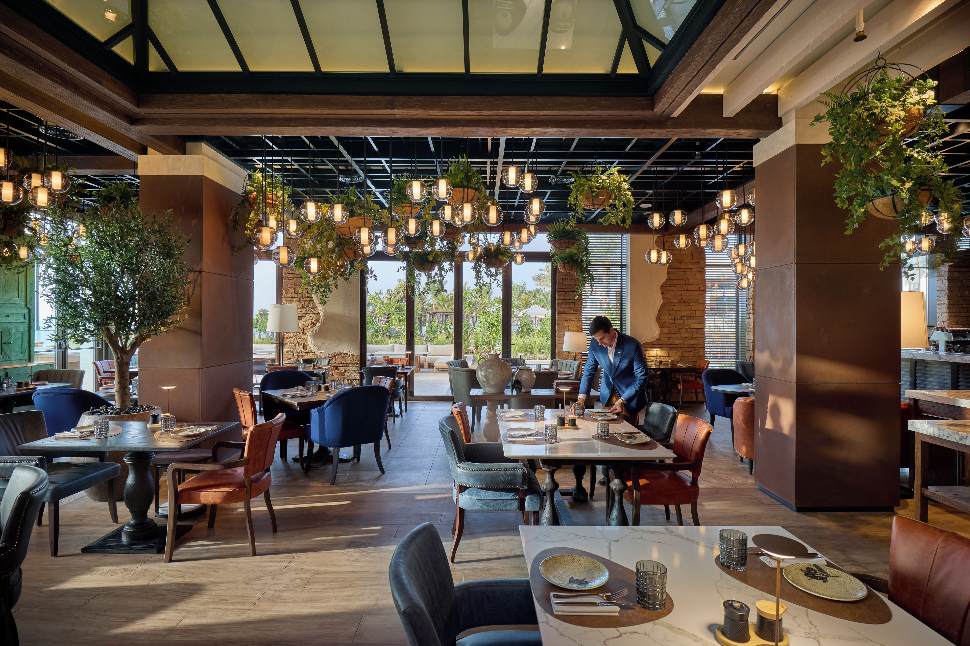 Un restaurante con varias mesas y sillas, plantas colgantes y una persona sirviendo comida.