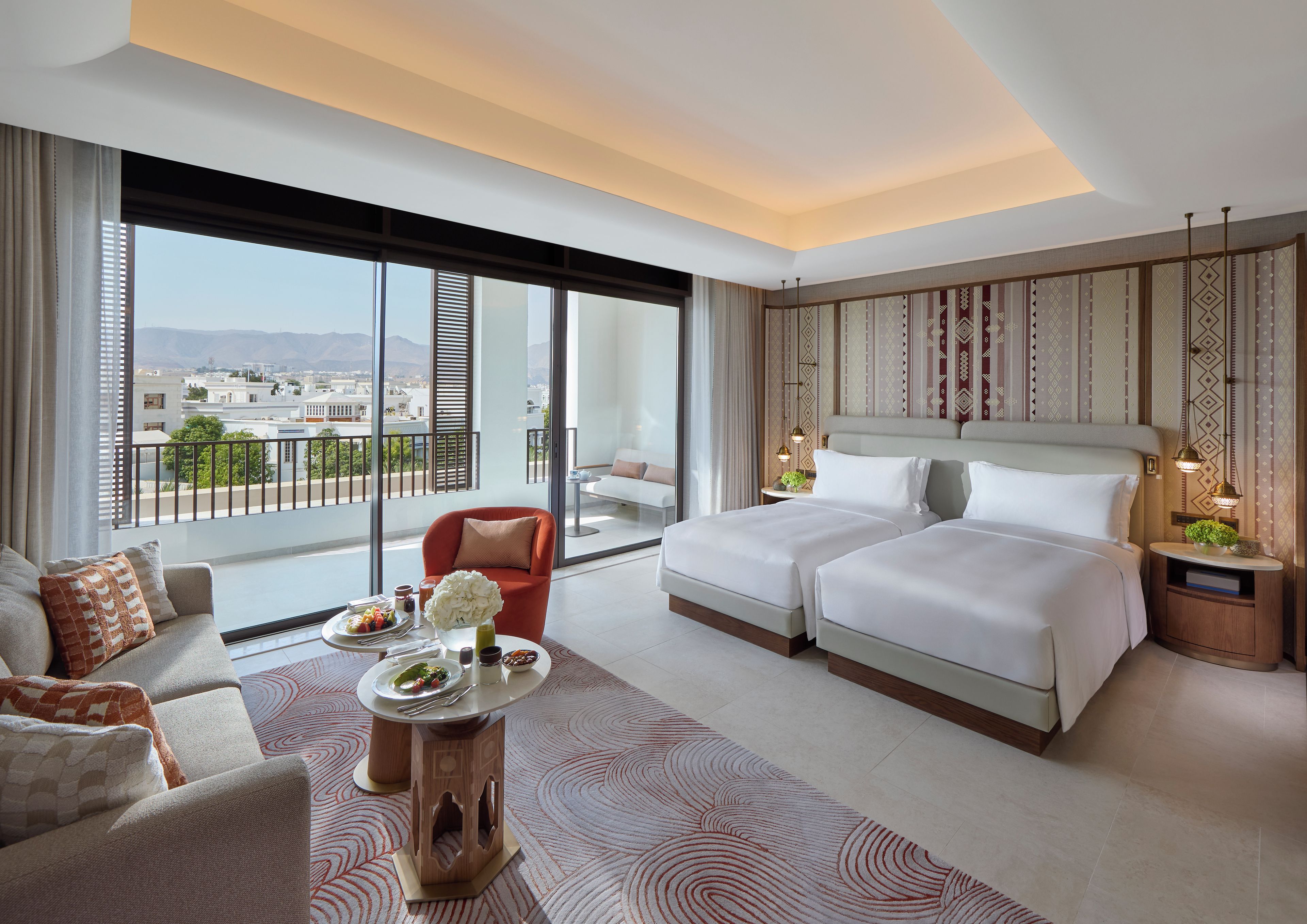Una habitación de hotel moderna con una cama grande, un sofá, una mesa de comedor con un florero y vistas a la ciudad a través de amplias ventanas.
