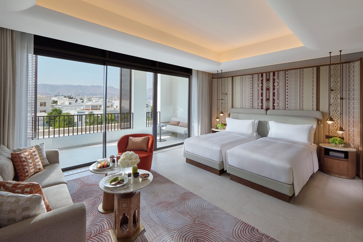 A modern hotel room with a large bed, a sofa, a dining table with a vase of flowers, and a view of the city through large windows.