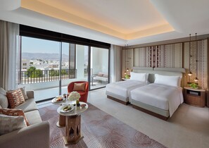 A modern hotel room with a large bed, a sofa, a dining table with a vase of flowers, and a view of the city through large windows. - Mandarin Oriental Muscat (Muscat)