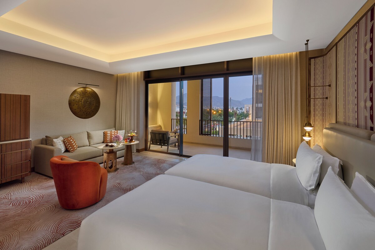 A hotel room with a large bed, a sofa, a small table with a vase of flowers, and a balcony with a view of the city.