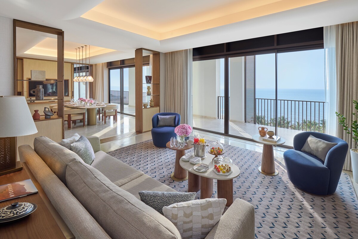 A modern hotel room with a sofa, armchairs, a dining table, and a balcony with a view.