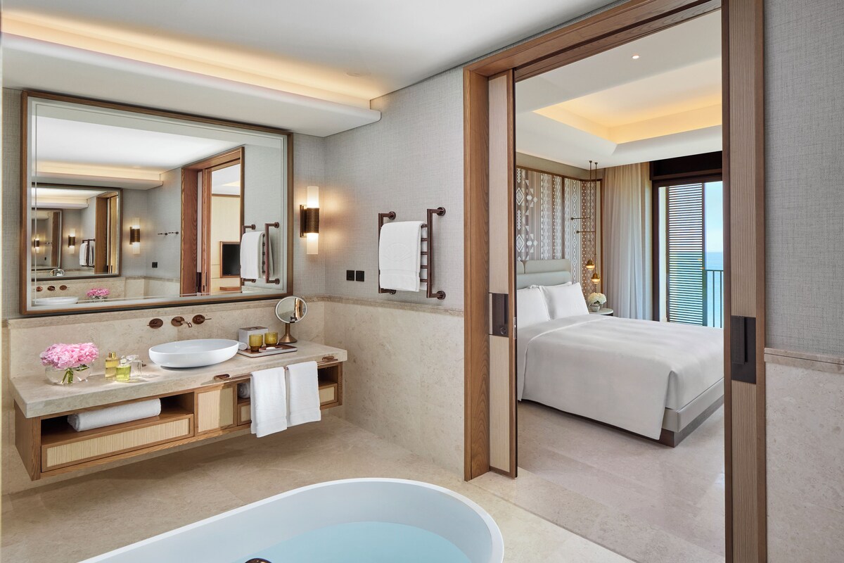 A modern bathroom with a freestanding bathtub, a large mirror, a vanity with a sink and towel rack, and a view of a bedroom through an open door.