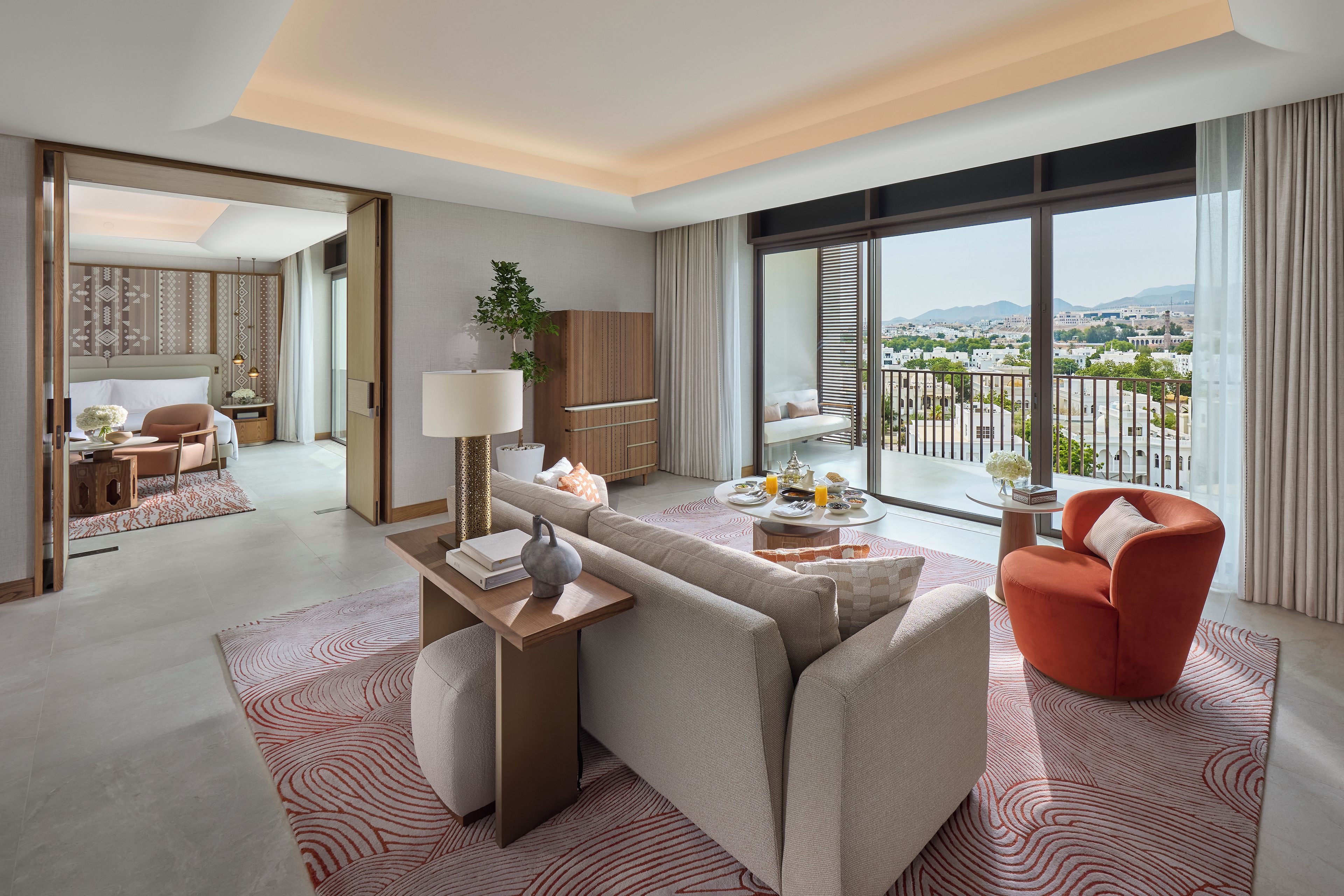 Una habitación de hotel moderna con balcón, sofá, sillón rojo, mesa de comedor y vistas a la ciudad.