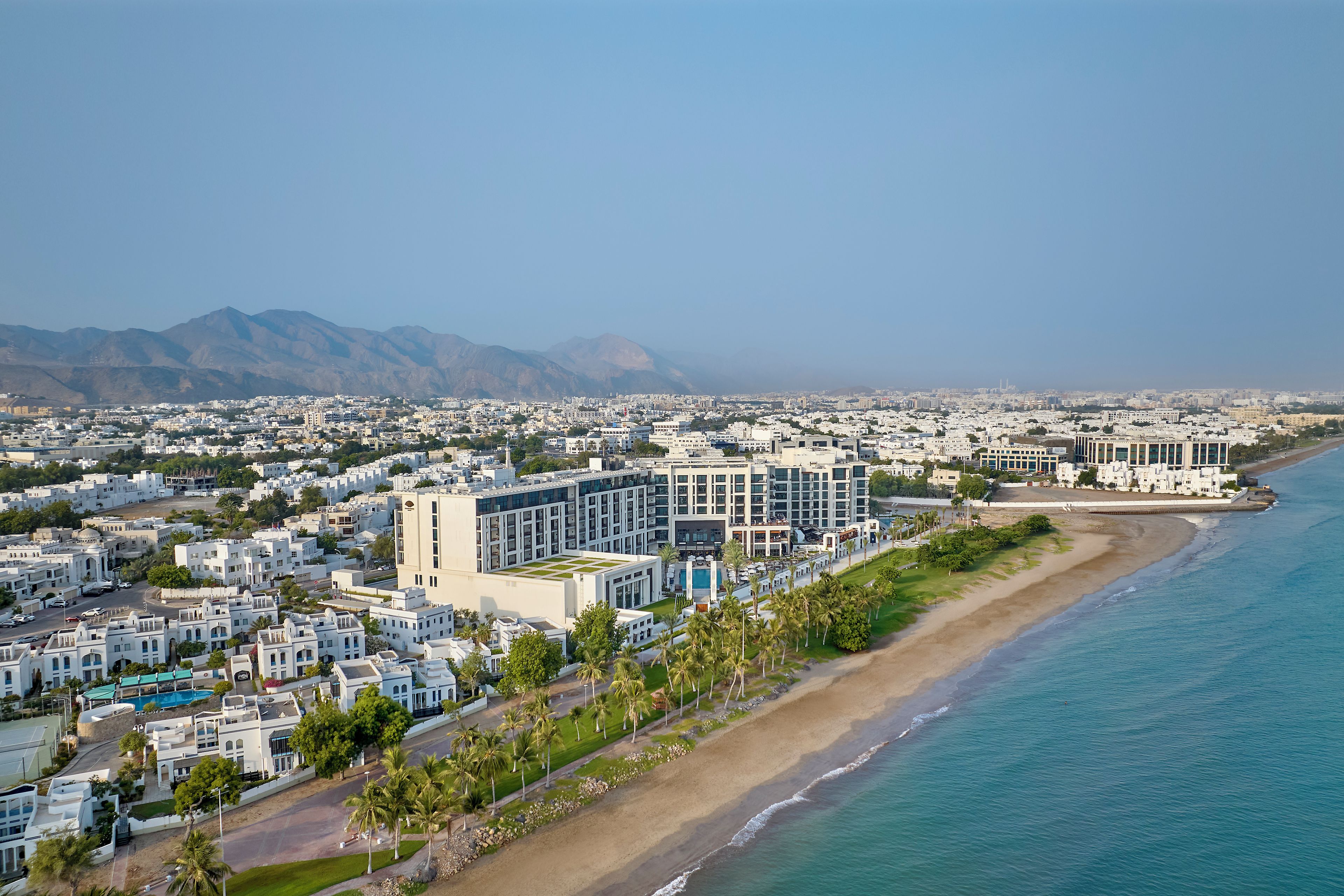 Un paisaje urbano costero con edificios residenciales, una playa y montañas al fondo.