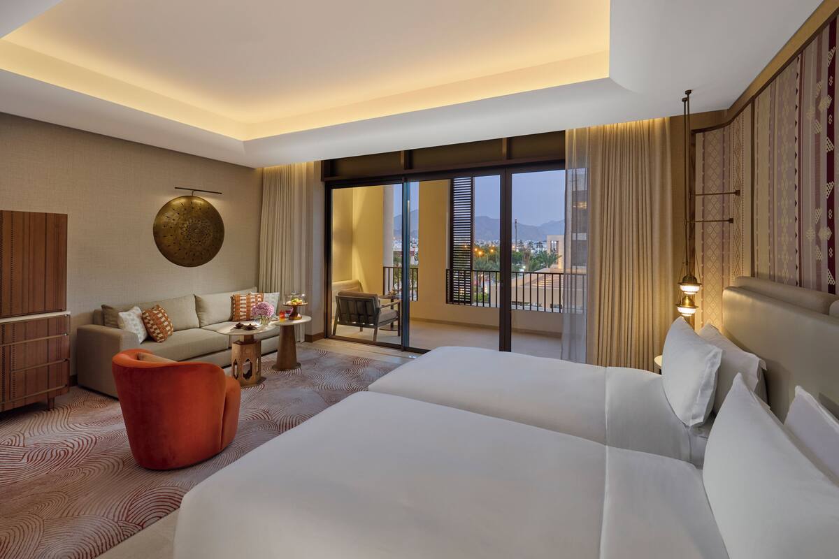 Habitación de hotel con una cama grande, un sofá, una mesita con un jarrón de flores y un balcón con vista a la ciudad.