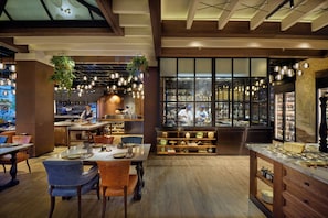 Interior de un restaurante con pisos de madera, plantas colgantes y vista a la cocina a través de amplios ventanales.