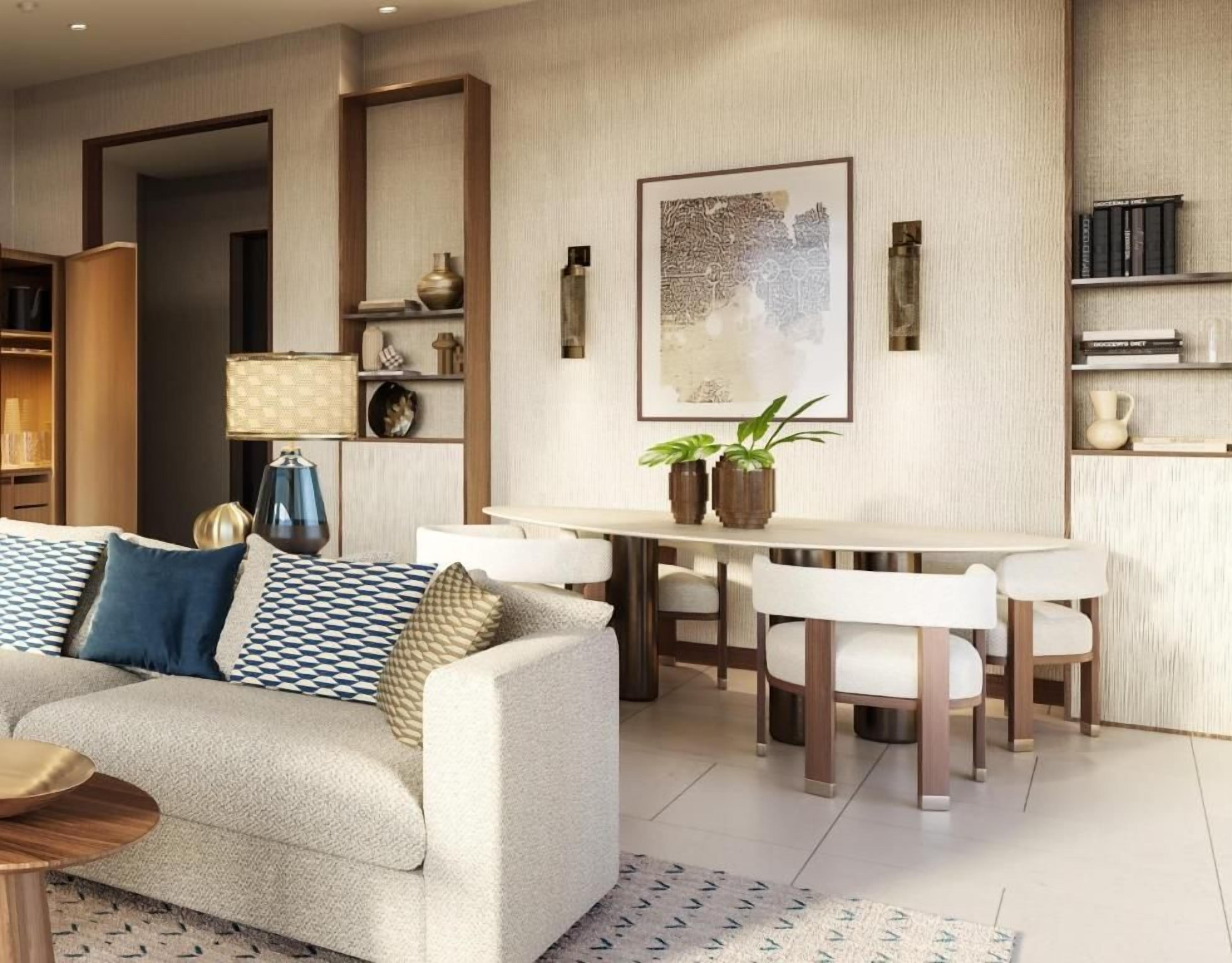 A modern living room with a sofa, dining table, and chairs. There is a bookshelf, a lamp, and a framed artwork on the wall.