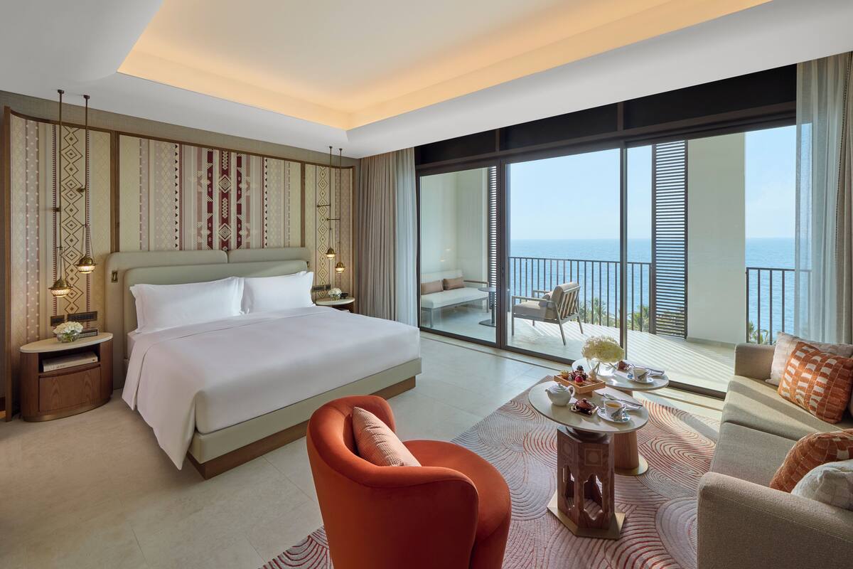 Habitación de hotel con una cama grande, un sofá, una mesa de comedor y un balcón con vista al mar.