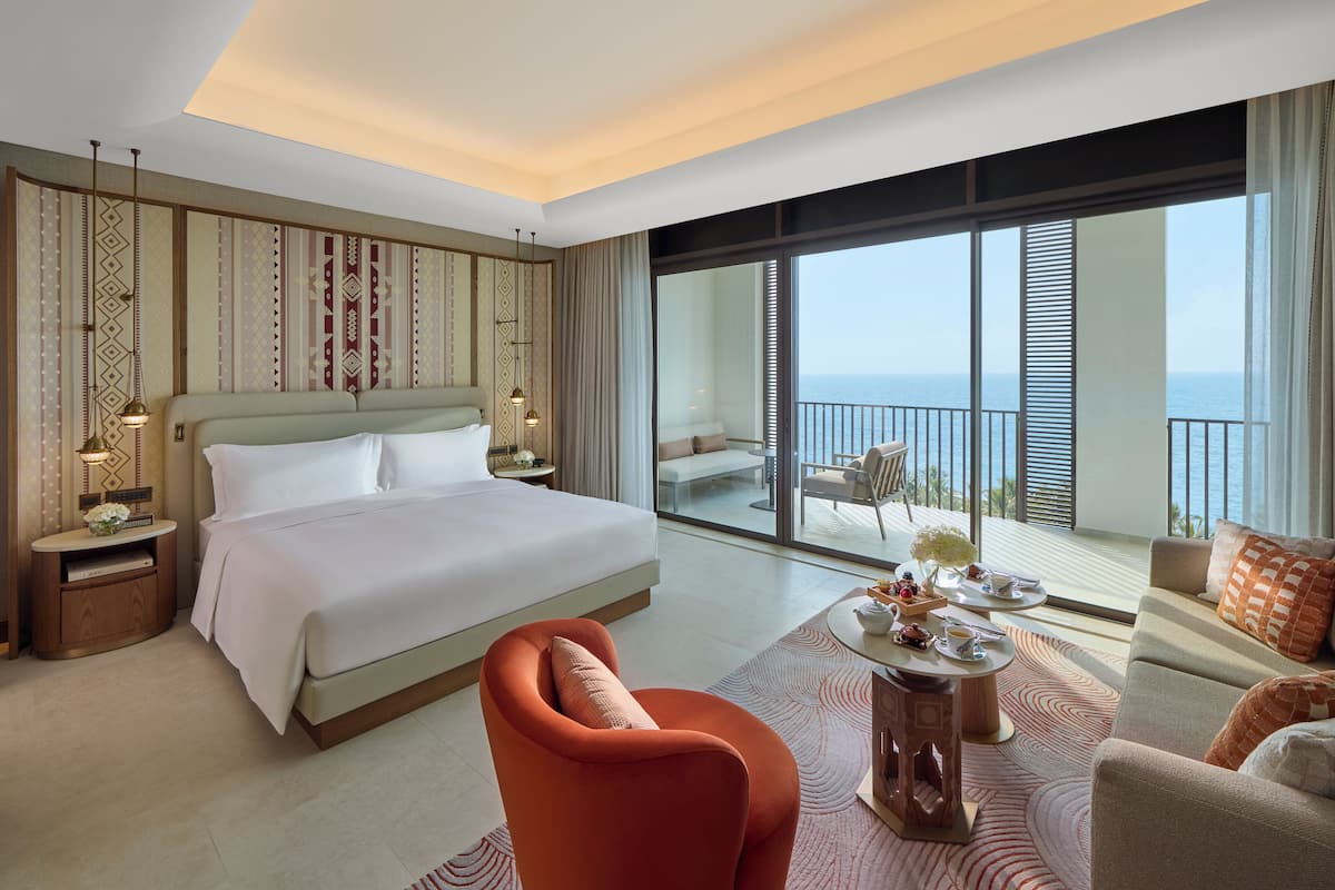 A hotel room with a large bed, a sofa, a dining table, and a balcony with a view of the sea.