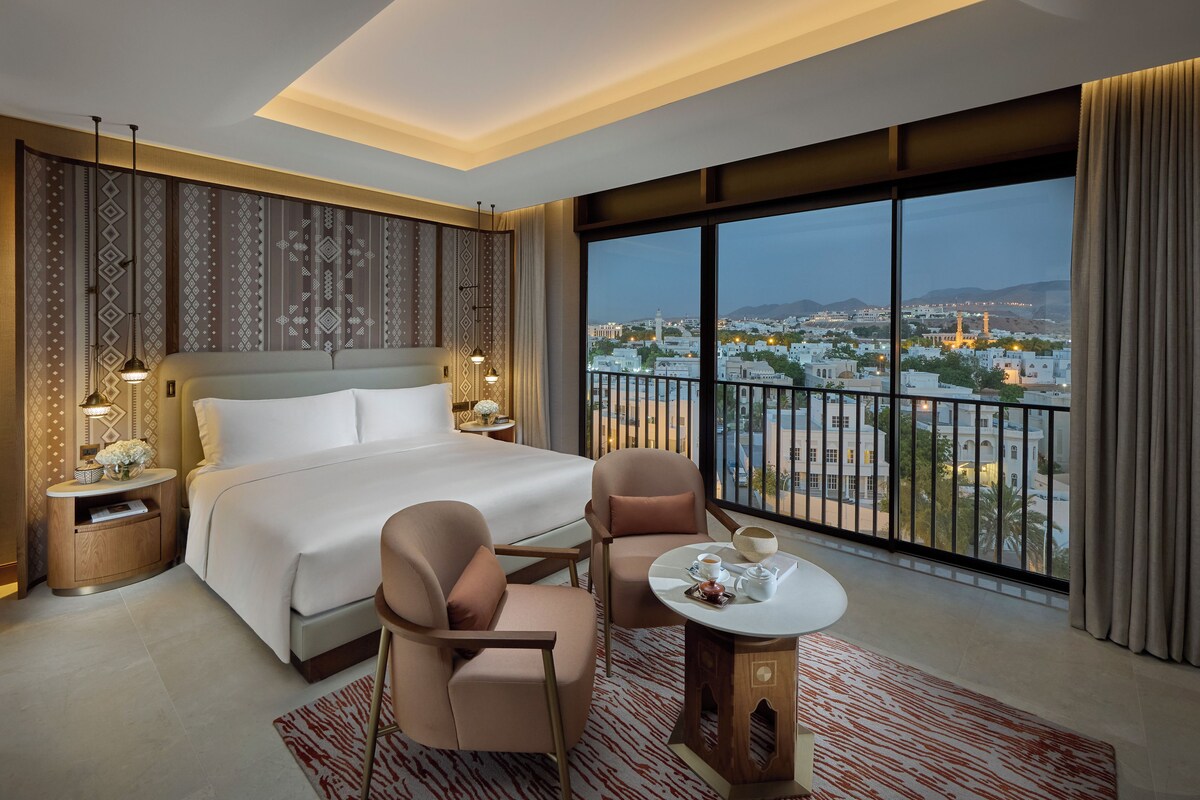 A hotel room with a large bed, two bedside tables, a chair, a small table with a cup and saucer, and a view of a cityscape through large windows.