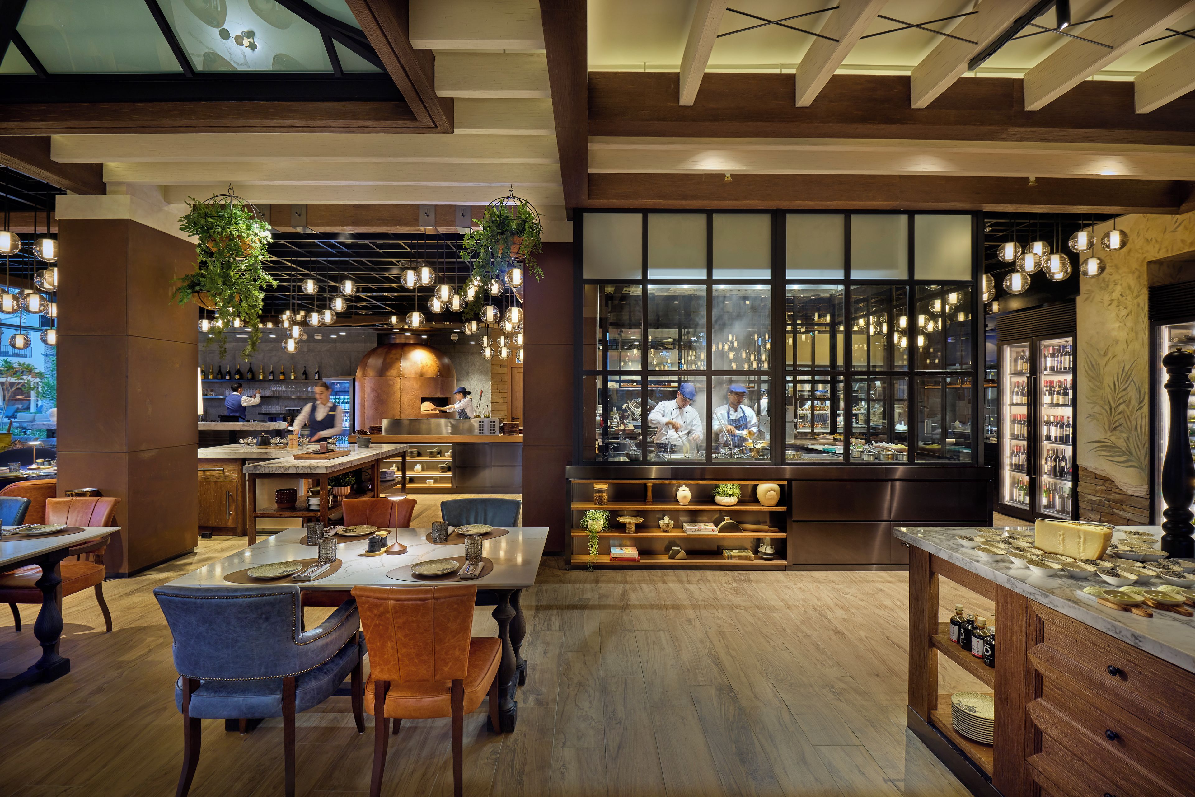 A restaurant interior with wooden floors, hanging plants, and a view of the kitchen through large windows.