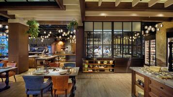 A restaurant interior with wooden floors, hanging plants, and a view of the kitchen through large windows.