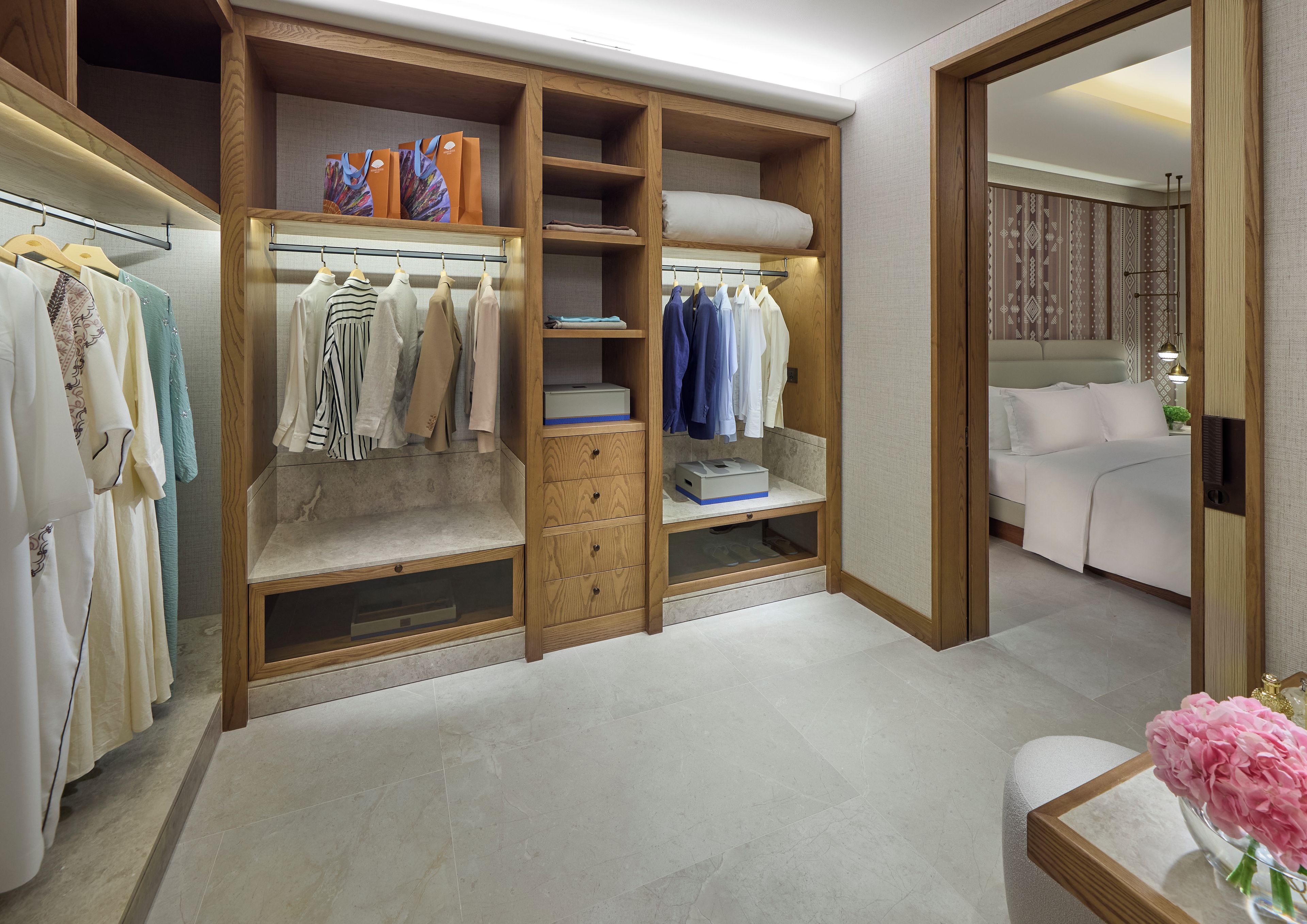 Un vestidor con estantes y cajones múltiples, una cama con ropa de cama blanca y una mesita con un florero.