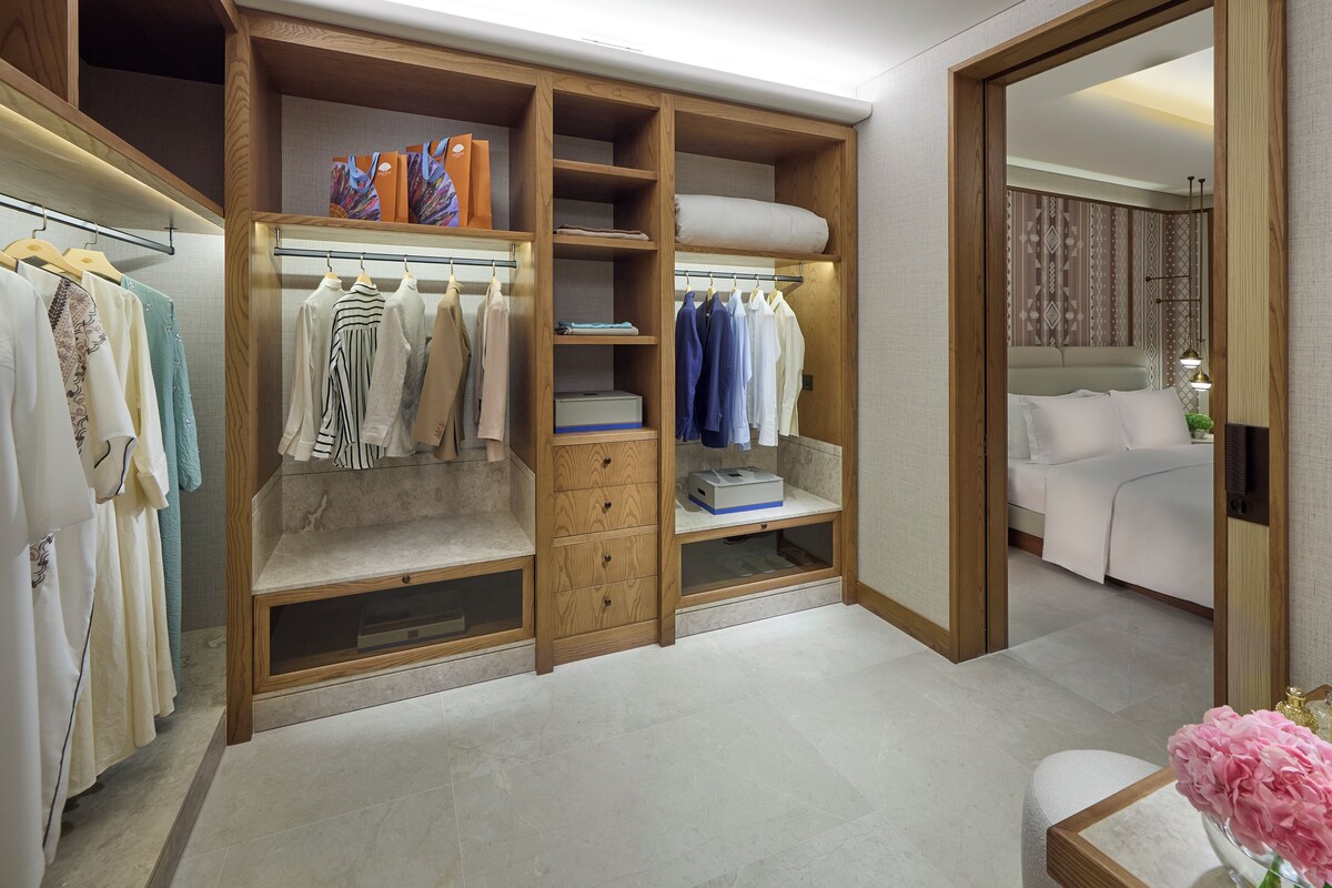 A walk-in closet with multiple shelves and drawers, a bed with white bedding, and a small table with a vase of flowers.