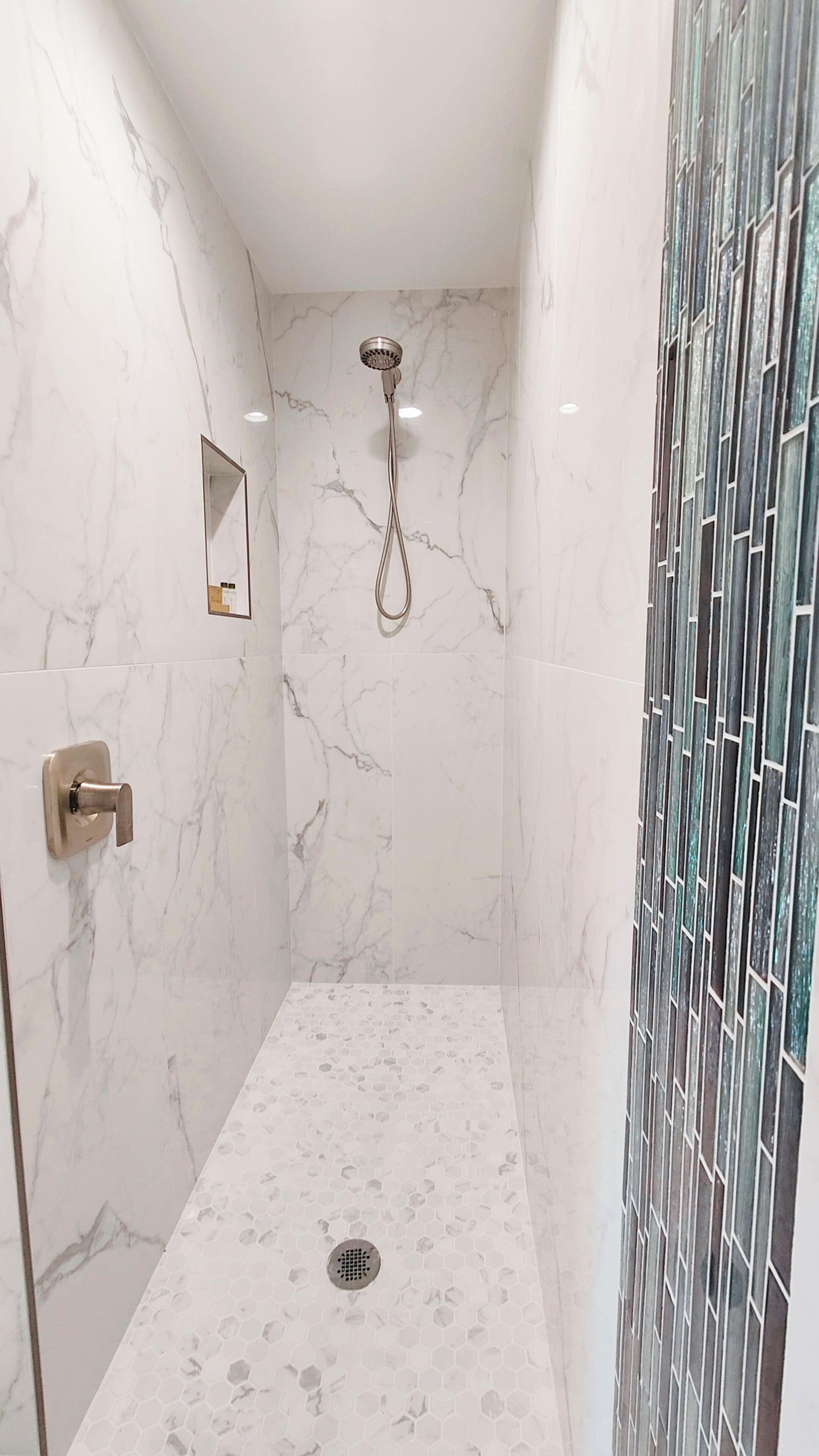 Un baño moderno con paredes de mármol, una ducha con cabezal de ducha manual y una puerta de vidrio con un diseño geométrico.