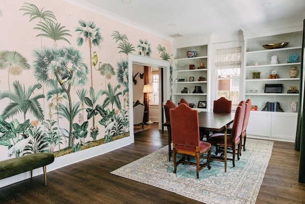 A dining area with a tropical mural, wooden dining table, red upholstered chairs, and a patterned rug.