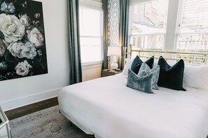 A neatly made bed with white linens and multiple pillows, a large floral artwork on the wall, and a window with curtains.