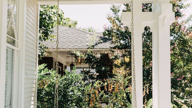 A porch with a swing chair, white columns, and a view of a house and garden. - Midtown Manor by Pettigrew Adventures (Memphis)