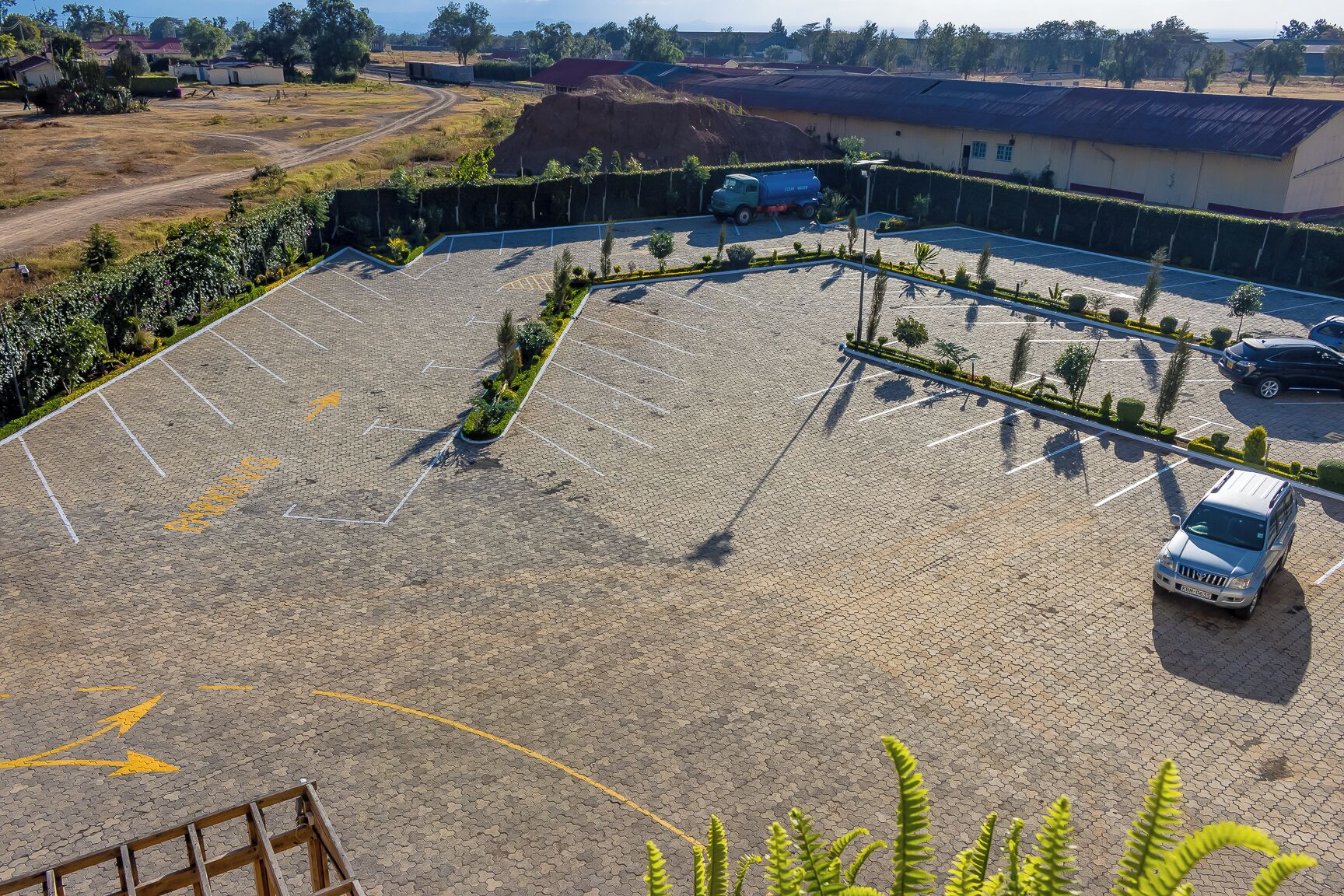 Vista aérea de um estacionamento com alguns veículos, cercado por vegetação e edifícios.