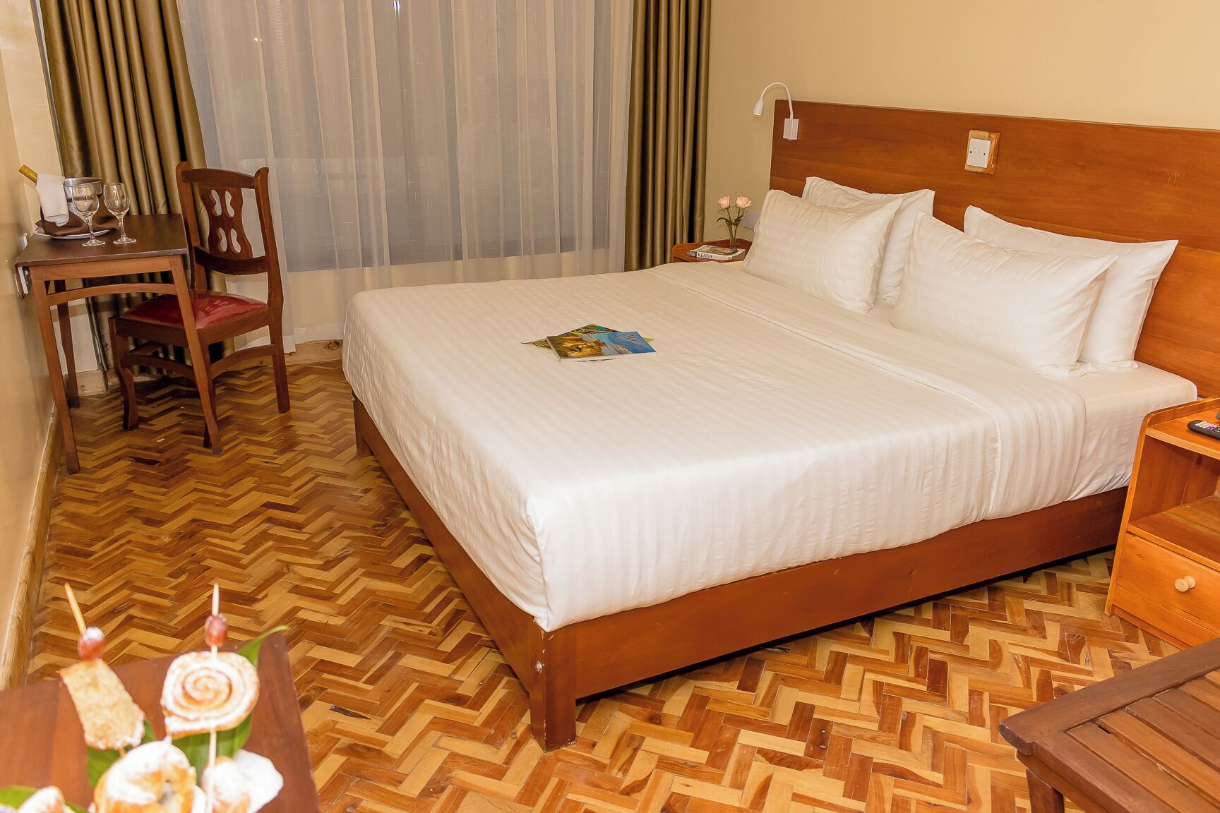 Habitación de hotel con una cama grande, cabecero de madera, una silla de madera, una mesita con un libro y un control remoto, y suelo de madera con un patrón de espiga.