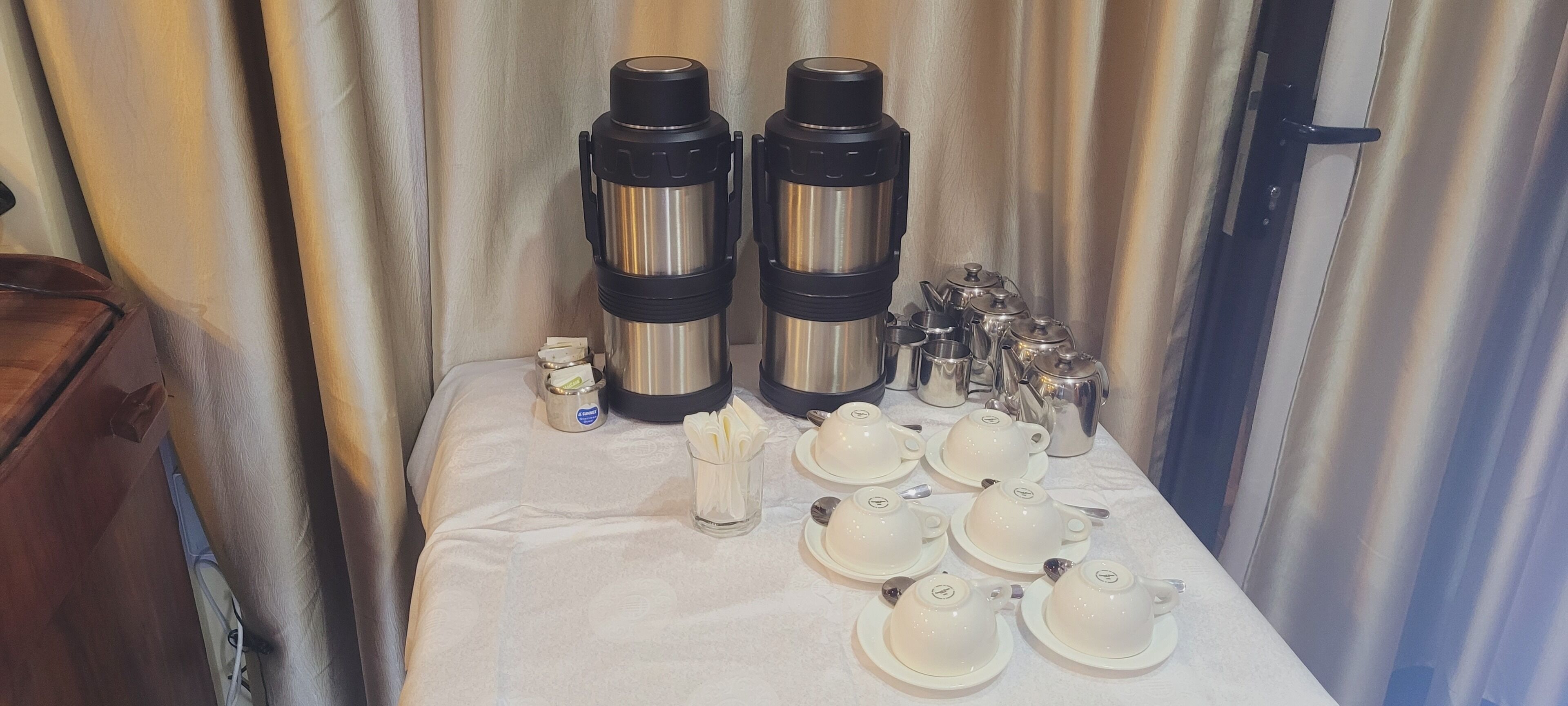 A table set with two stainless steel thermos flasks, a small container, and multiple teacups and saucers.