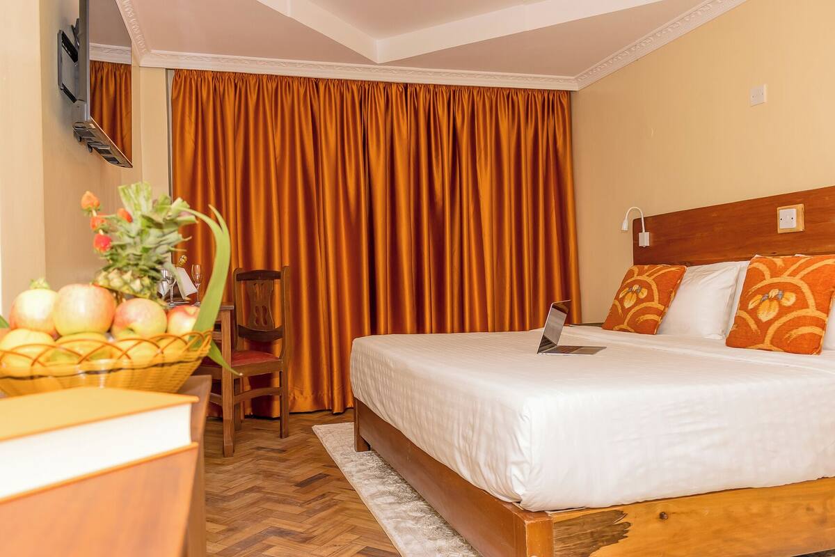 Una habitación de hotel con una cama, un escritorio con una computadora portátil, una silla, una cesta de fruta y una cortina.