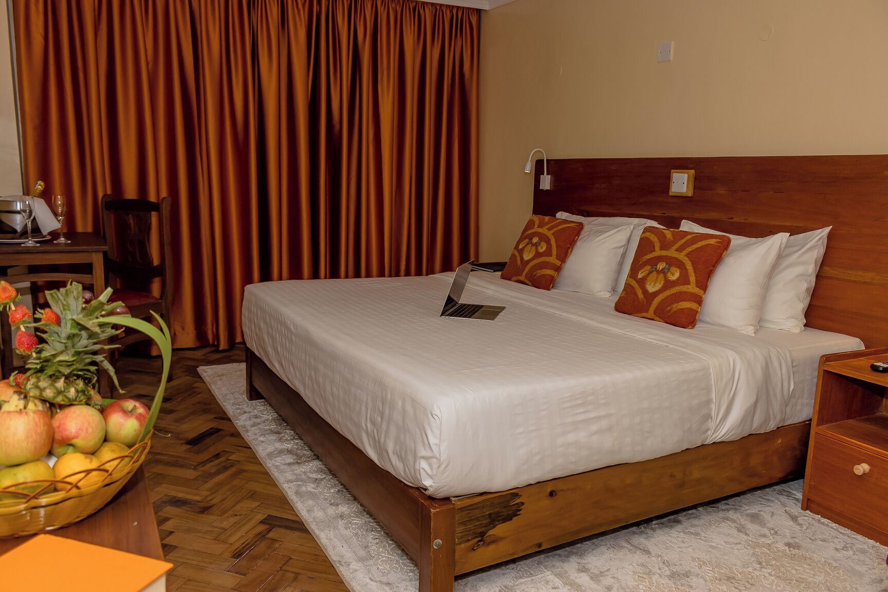 A hotel room with a wooden bed, white bedding, a laptop on the bed, a fruit basket, and a wooden nightstand.
