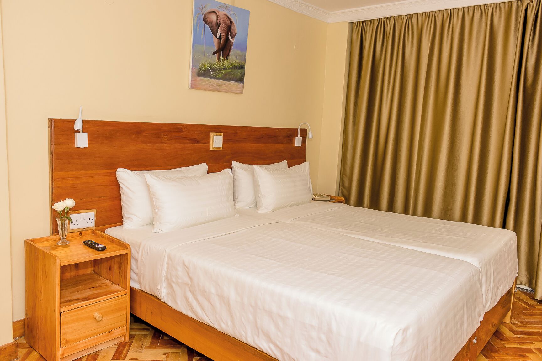 Ein Hotelzimmer mit einem hölzernen Kopfteil, einem großen Bett mit weißen Leinentüchern, einem Nachttisch mit Blumen in einer Vase, einer Fernbedienung und einem Elefantenbild an der Wand.
