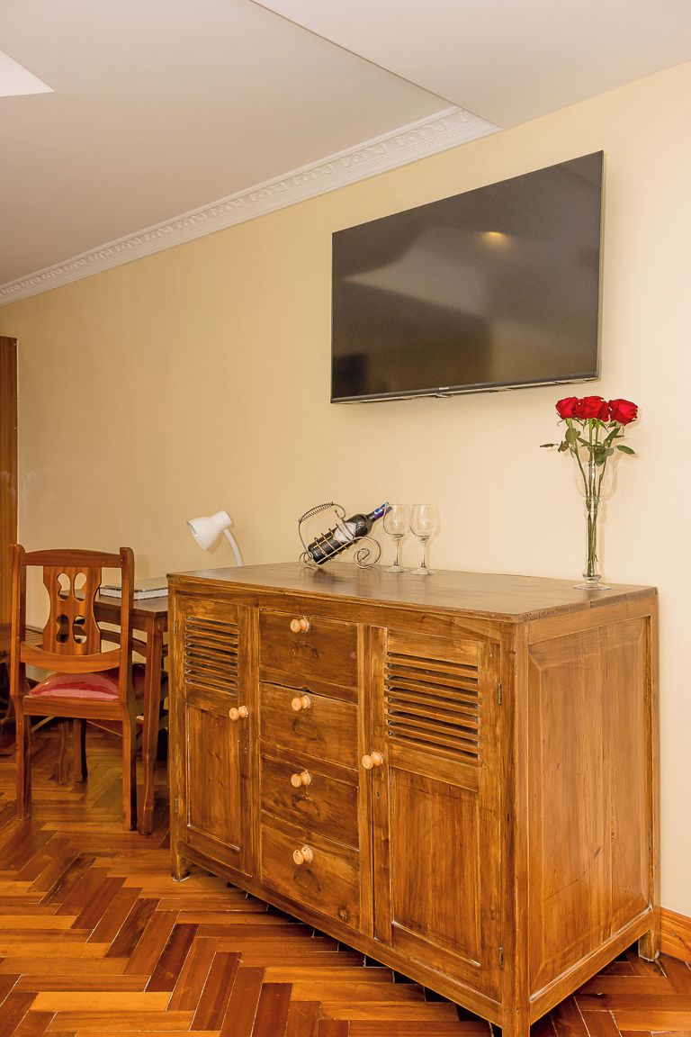 A wooden cabinet with drawers, a television mounted on the wall, a vase with red roses, and a chair.