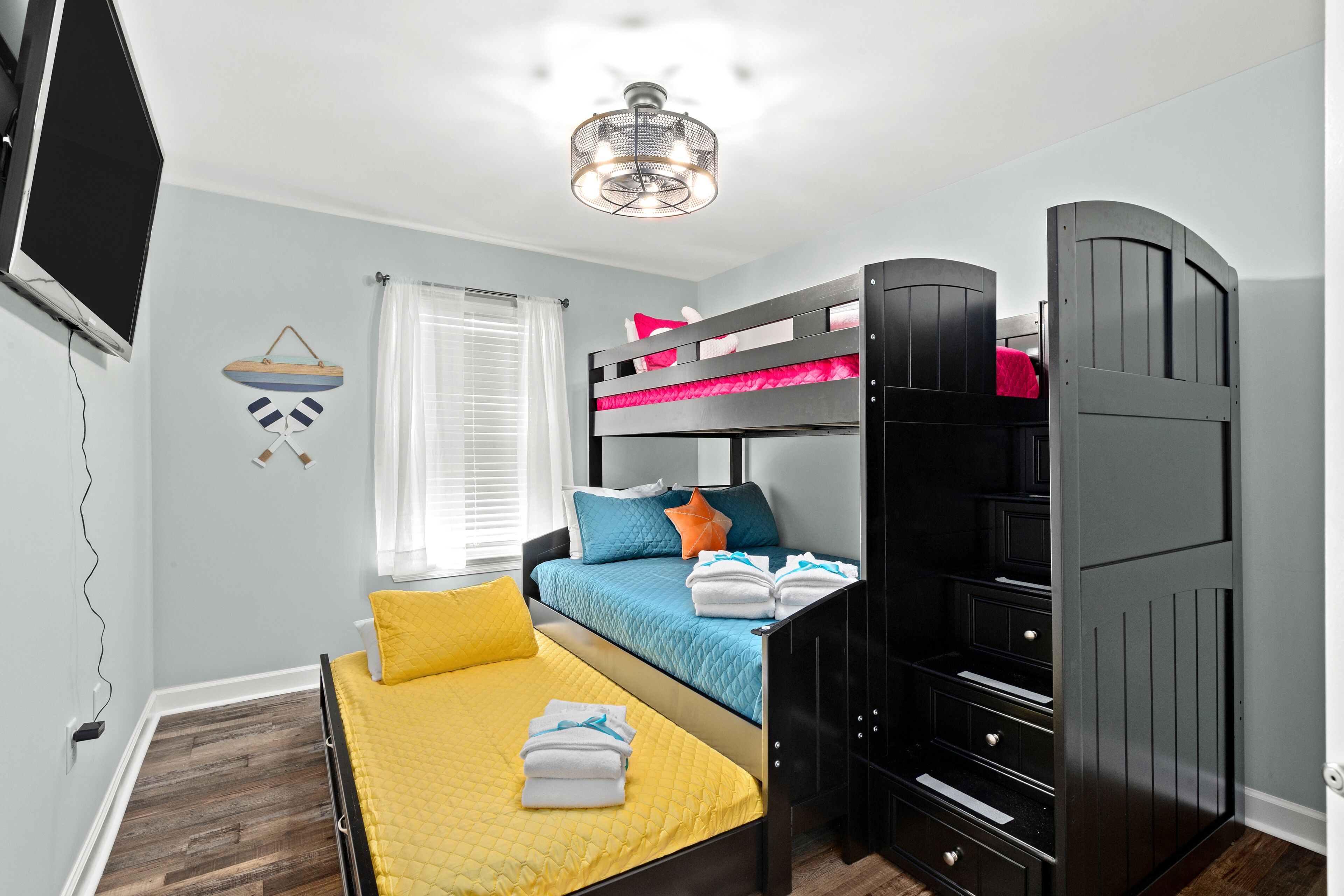Une chambre avec un lit superposé, un lit simple avec une literie jaune, un ventilateur de plafond et un téléviseur fixé au mur.