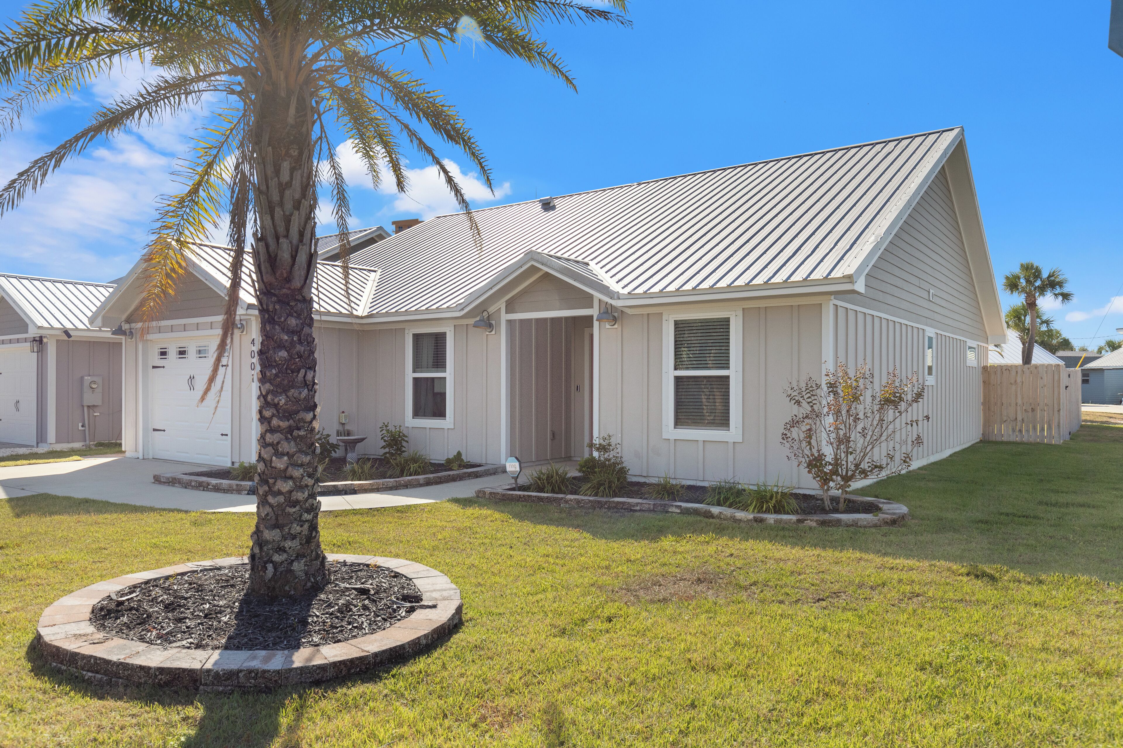 A single-story house with a sloped metal roof, a front porch, and a small garden with a palm tree.