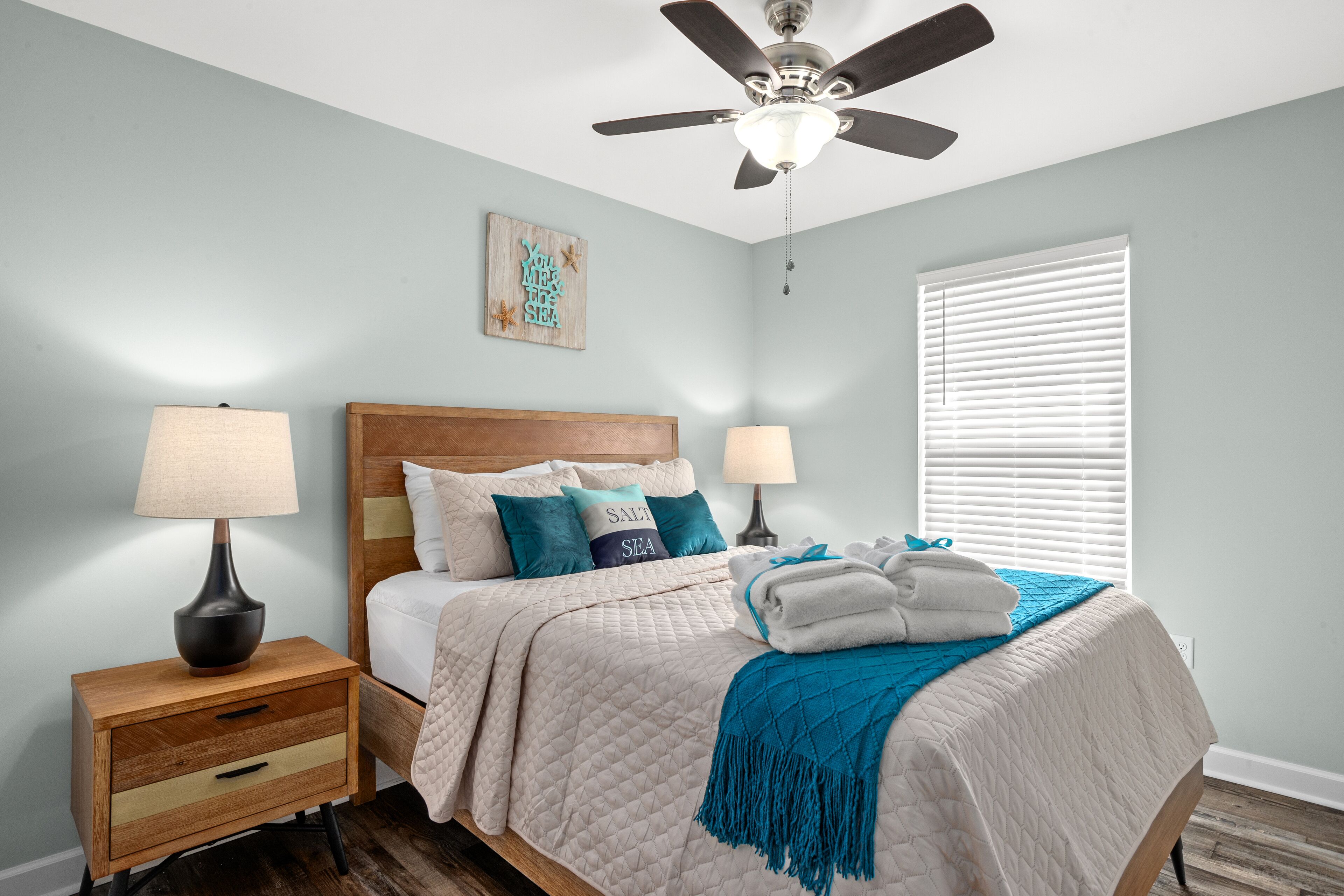 A bedroom with a bed, two bedside tables with lamps, a ceiling fan, a window with blinds, and a wall decoration.