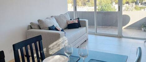 A modern living room with a sofa, dining table set with plates and wine glasses, and a balcony with a view of trees and buildings.