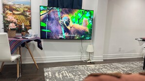 Un salón con un televisor de pantalla plana montado en la pared, una silla blanca, una mesita con un jarrón y una botella, y una lámpara.